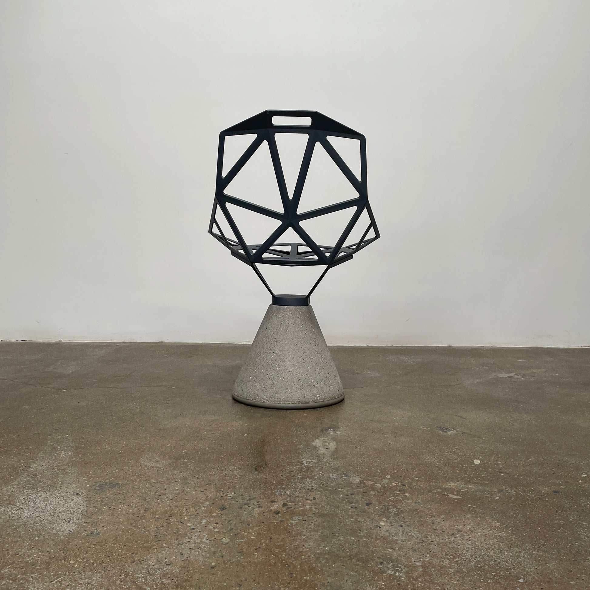 The Magis Grey Swivel Chair One With Concrete Base, designed by Konstantin Grcic, showcases a geometric open lattice design on a concrete base, sitting gracefully on a textured floor against a plain wall.