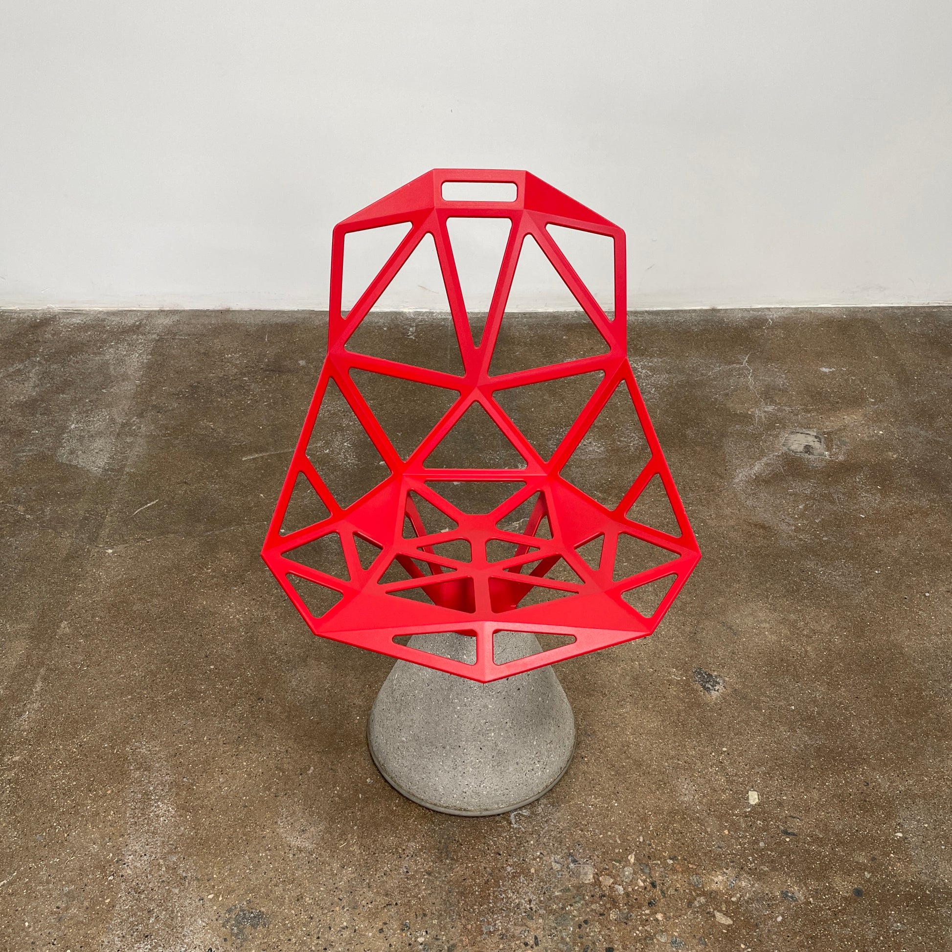 A Magis Red Chair One with a swivel base is positioned on a concrete foundation against a plain white background, reflecting the innovative style of Konstantin Grcic's designs.