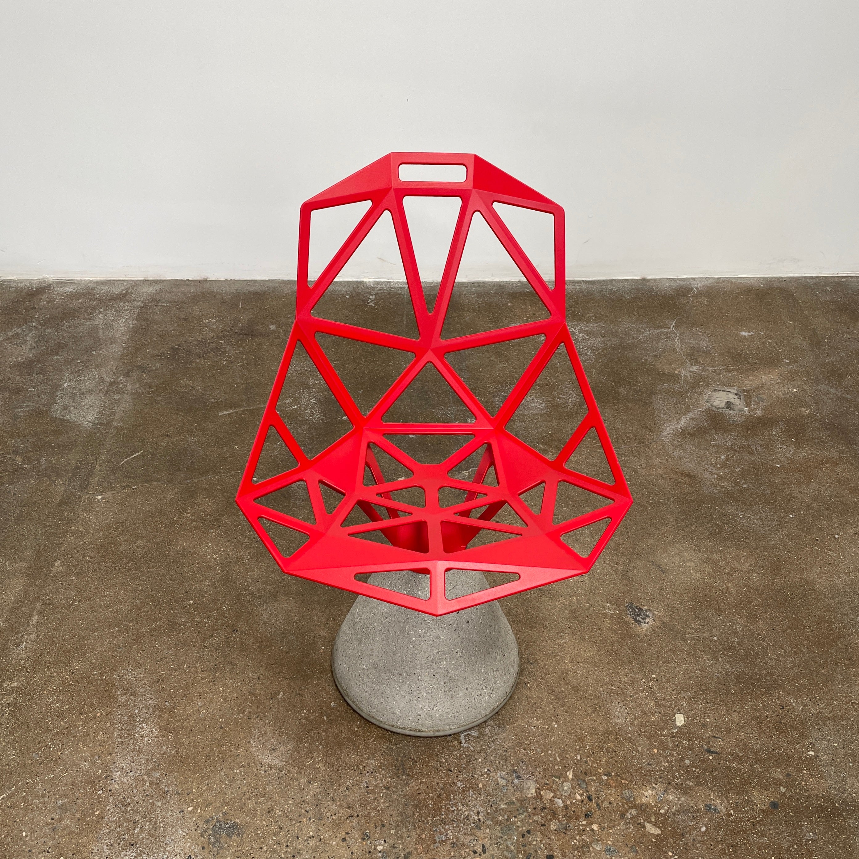 A Magis Red Chair One with a swivel base is positioned on a concrete foundation against a plain white background, reflecting the innovative style of Konstantin Grcic's designs.