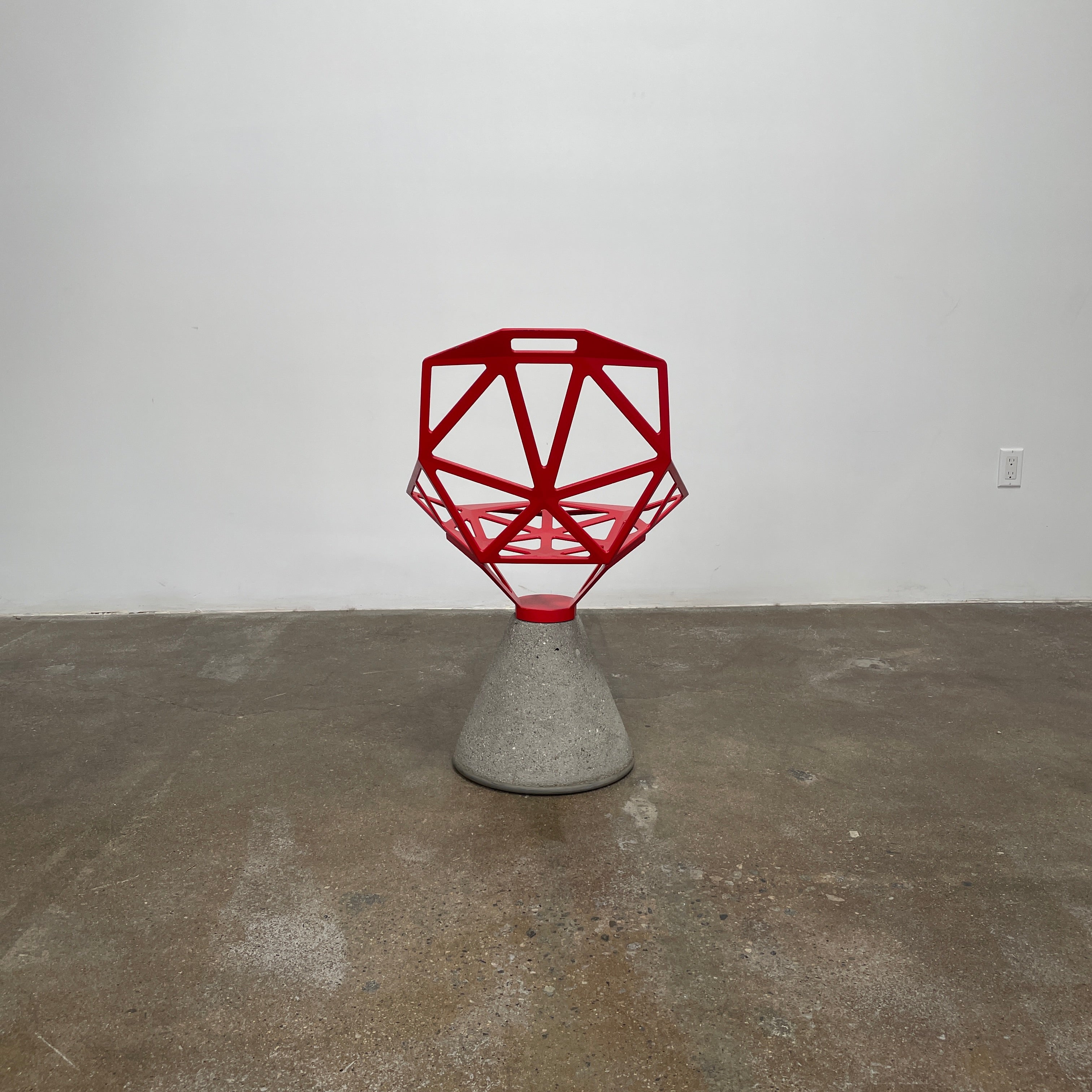 A Magis Red Chair One with a swivel base is positioned on a concrete foundation against a plain white background, reflecting the innovative style of Konstantin Grcic's designs.
