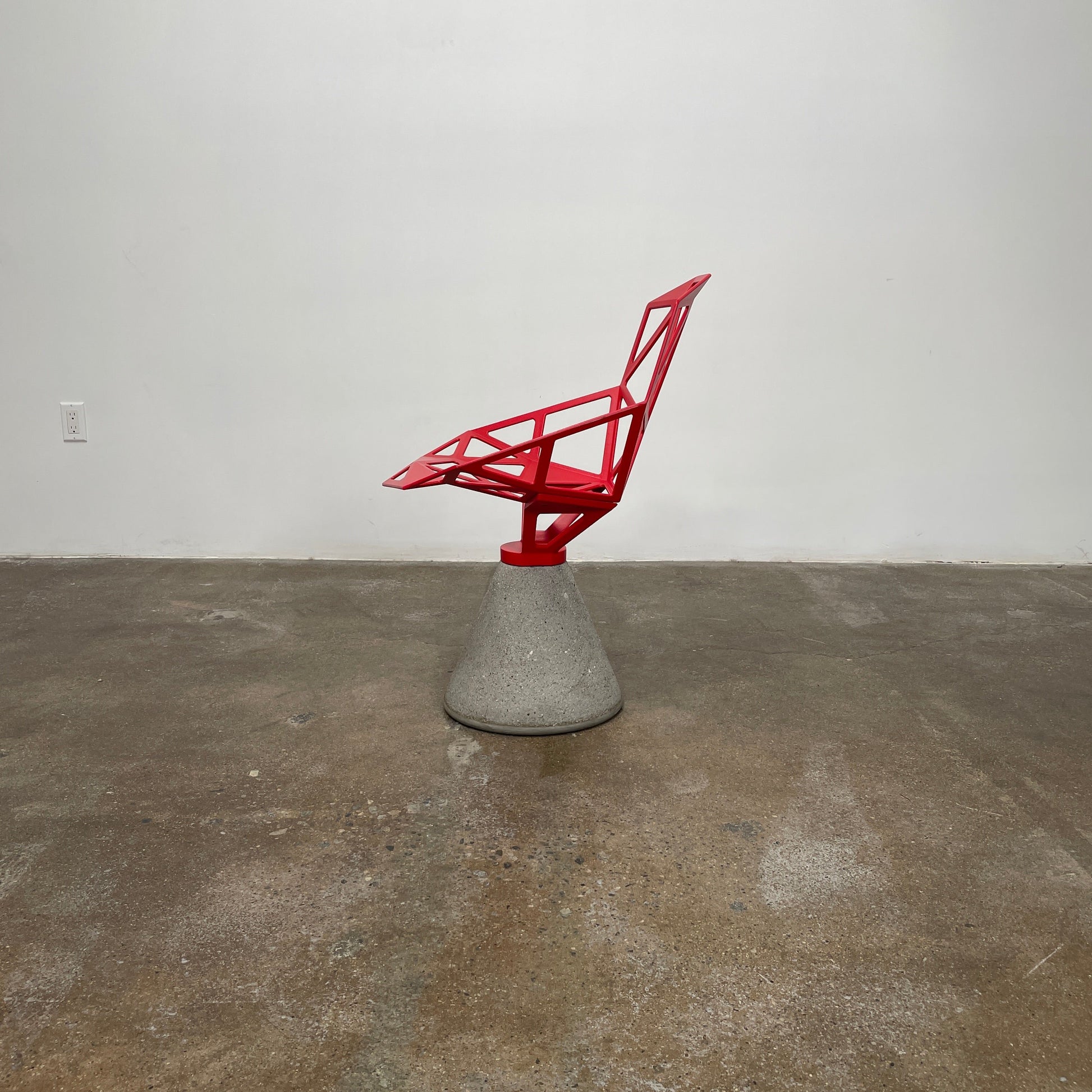 A Magis Red Chair One with a swivel base is positioned on a concrete foundation against a plain white background, reflecting the innovative style of Konstantin Grcic's designs.