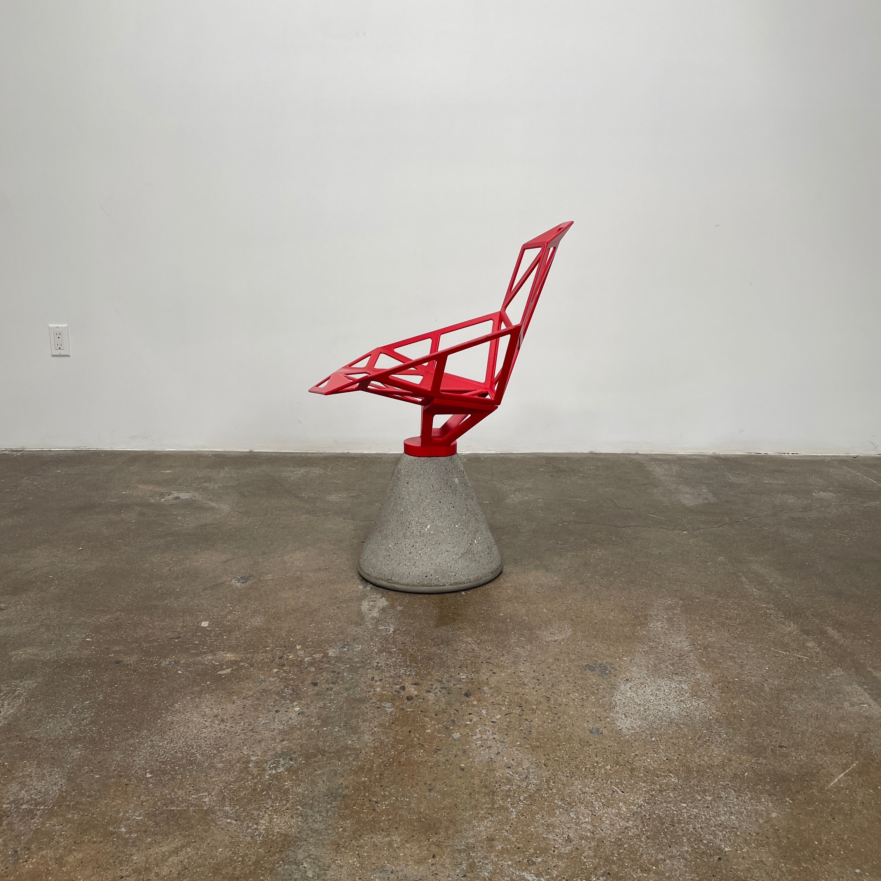 A Magis Red Chair One with a swivel base is positioned on a concrete foundation against a plain white background, reflecting the innovative style of Konstantin Grcic's designs.