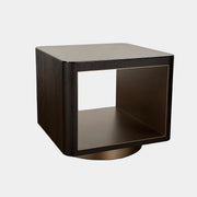 The Minotti Milton Side Table by Minotti features a modern, dark wooden cube design with an open center and a circular base, displayed on a polished concrete floor against a plain white wall.