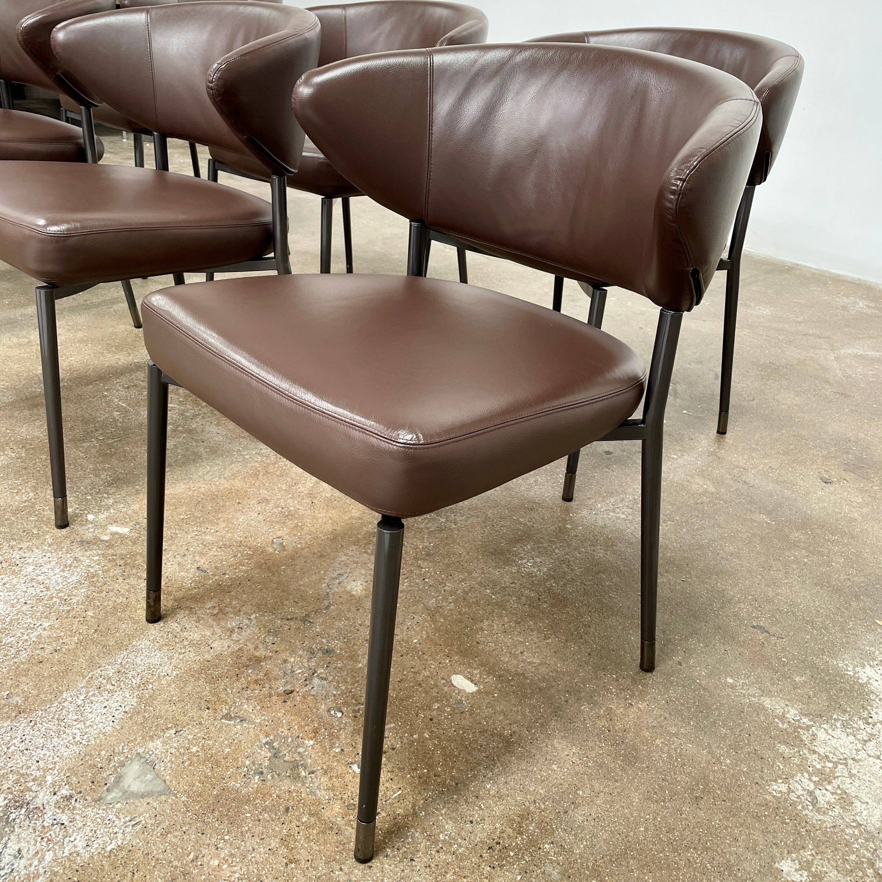 A set of six Minotti Mills Dining Chairs, featuring Scandinavian design with brown leather upholstery and black metal legs, is arranged in a staggered formation against a plain background.