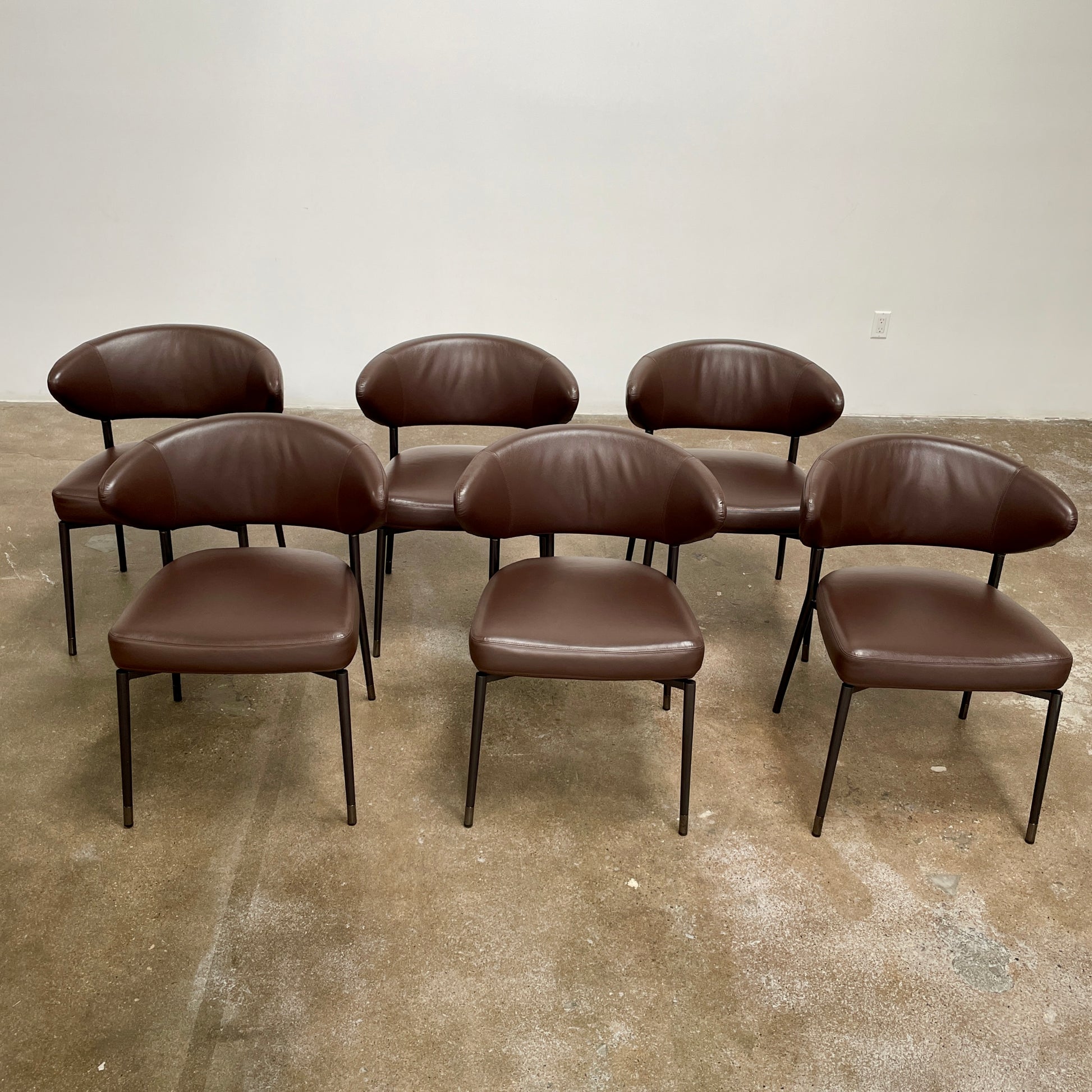 A set of six Minotti Mills Dining Chairs, featuring Scandinavian design with brown leather upholstery and black metal legs, is arranged in a staggered formation against a plain background.