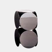 The Minotti Still Accent Table by Minotti features four large, circular, reflective metal discs stacked vertically for minimal elegance, shown on a concrete floor against a white wall.