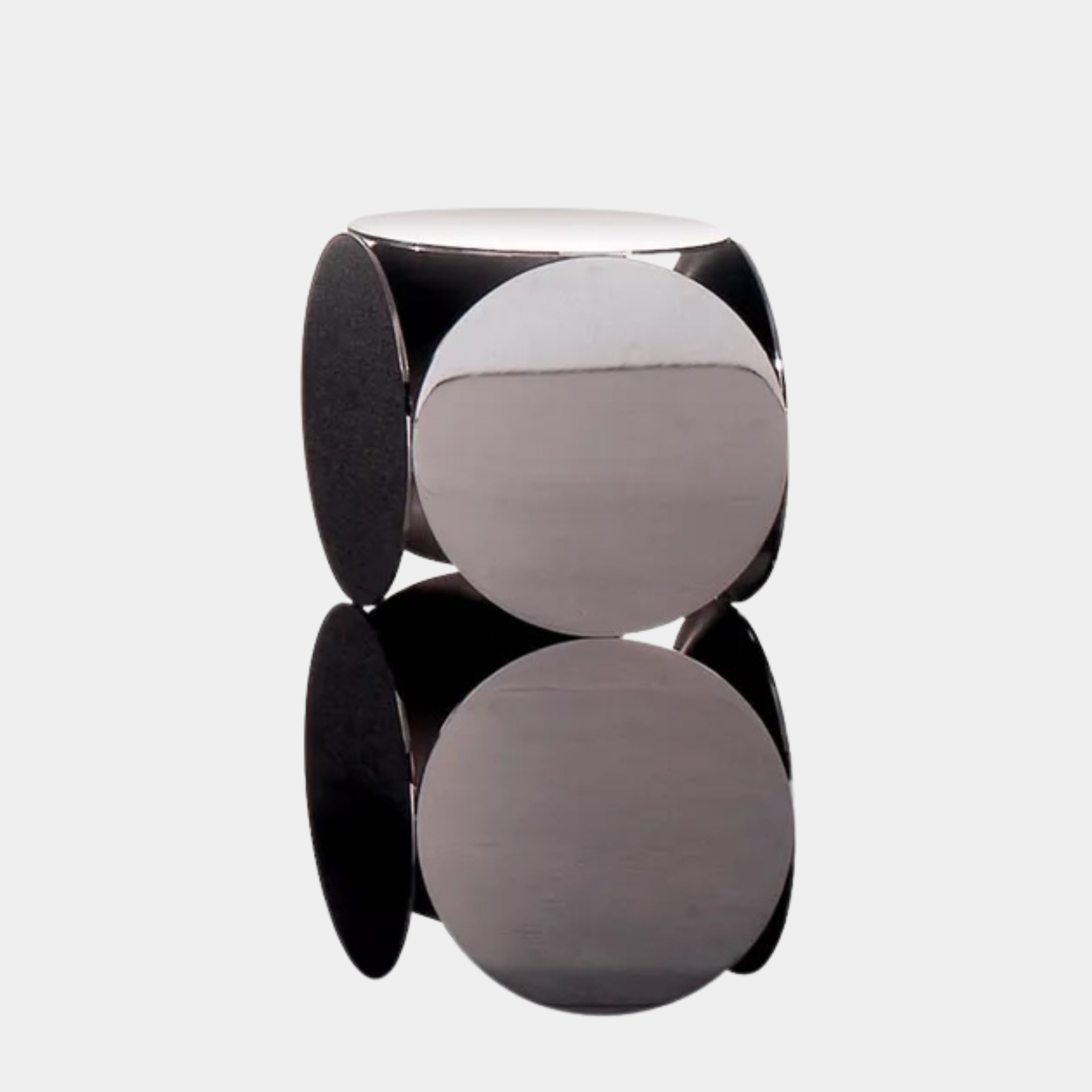 The Minotti Still Accent Table by Minotti features four large, circular, reflective metal discs stacked vertically for minimal elegance, shown on a concrete floor against a white wall.
