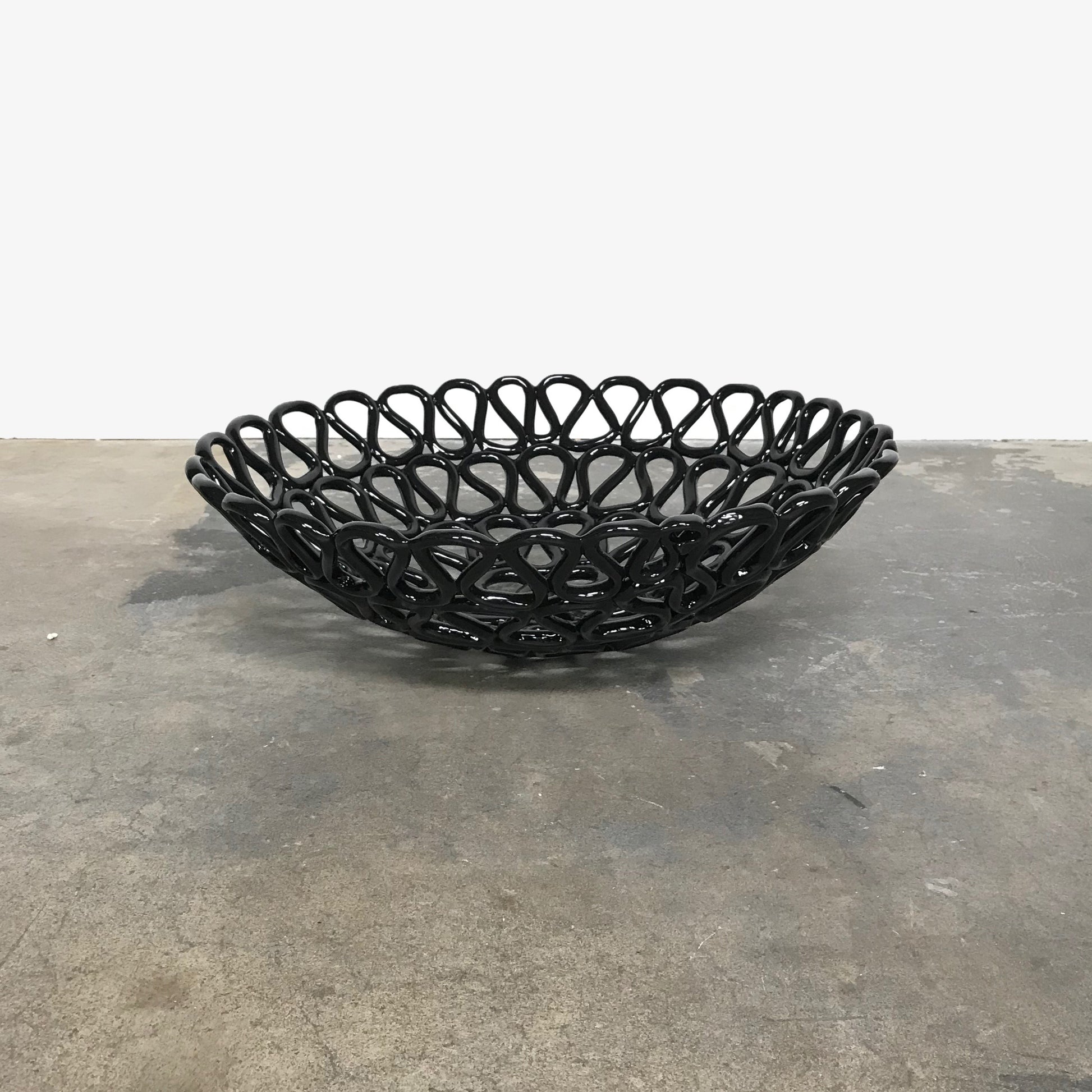 A Tom Dixon Fresh Fat Bowl in black with an intricate weave rests on a concrete floor, set against a simple white backdrop.
