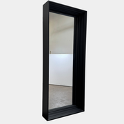 A person kneels in front of the Moooi Frame Mirror by Moooi—a large, rectangular mirror with a sleek black aluminum frame—while taking a photo. The mirror reflects furniture and industrial shelving within a well-lit room.