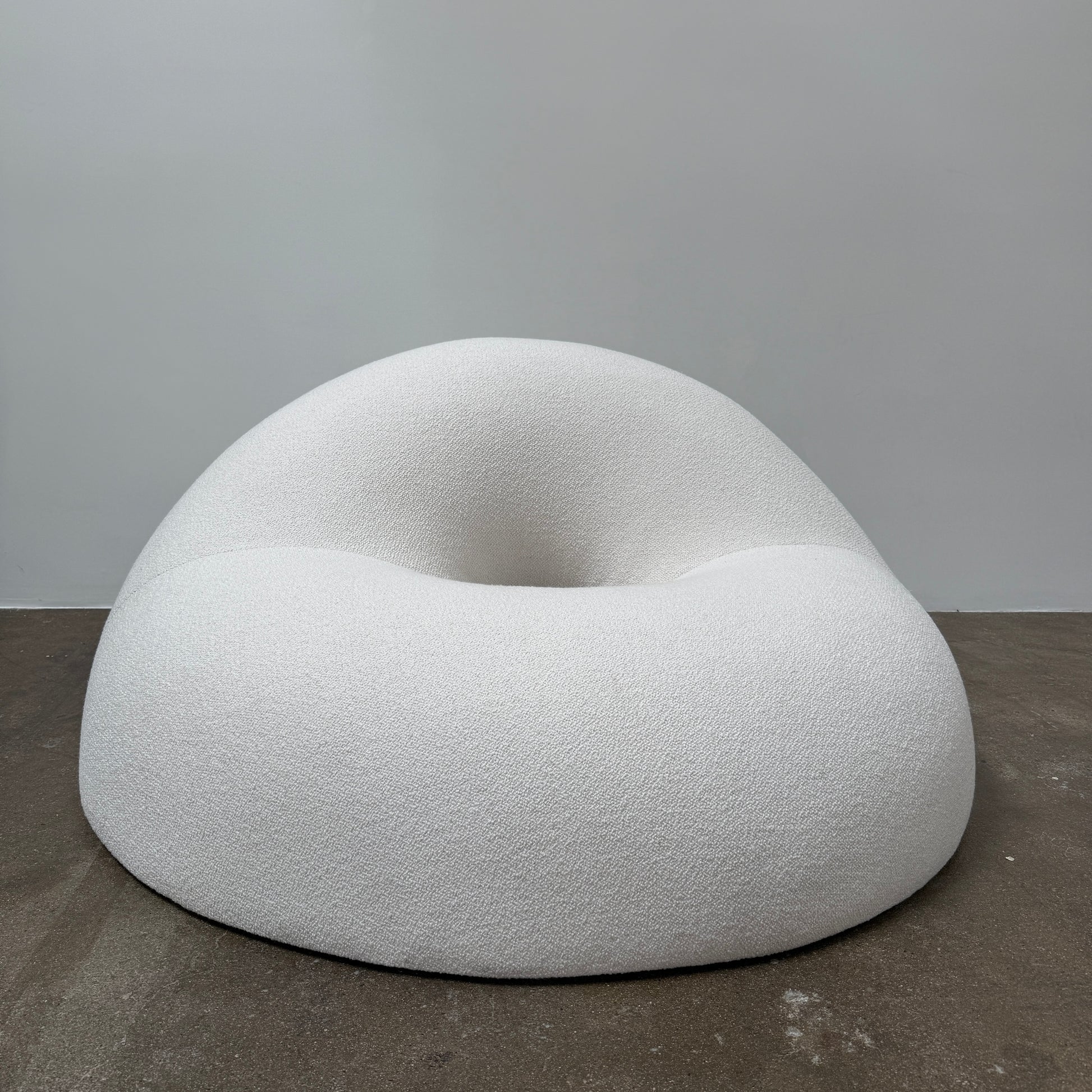 The Objects of Common Interest Tube Chair by Objects of Common Interest is a modern, light grey lounge chair with a donut-shaped design, rounded form, and central indentation, shown against a plain white background.