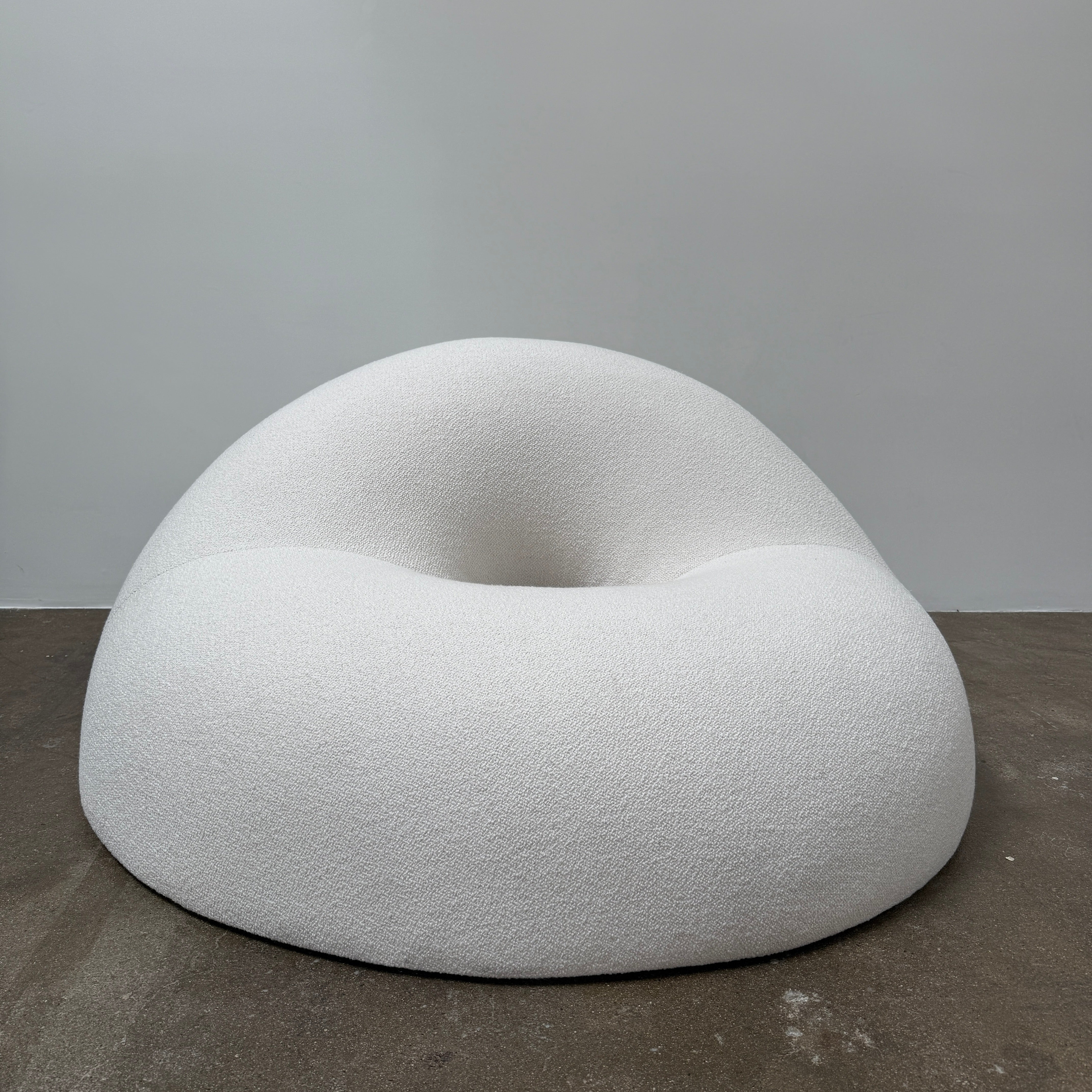 The Objects of Common Interest Tube Chair by Objects of Common Interest is a modern, light grey lounge chair with a donut-shaped design, rounded form, and central indentation, shown against a plain white background.