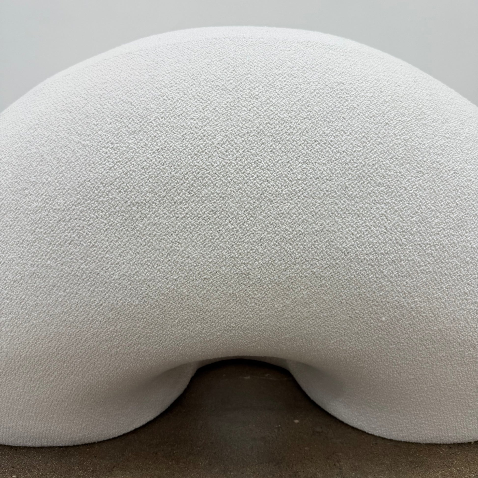 The Objects of Common Interest Tube Chair by Objects of Common Interest is a modern, light grey lounge chair with a donut-shaped design, rounded form, and central indentation, shown against a plain white background.