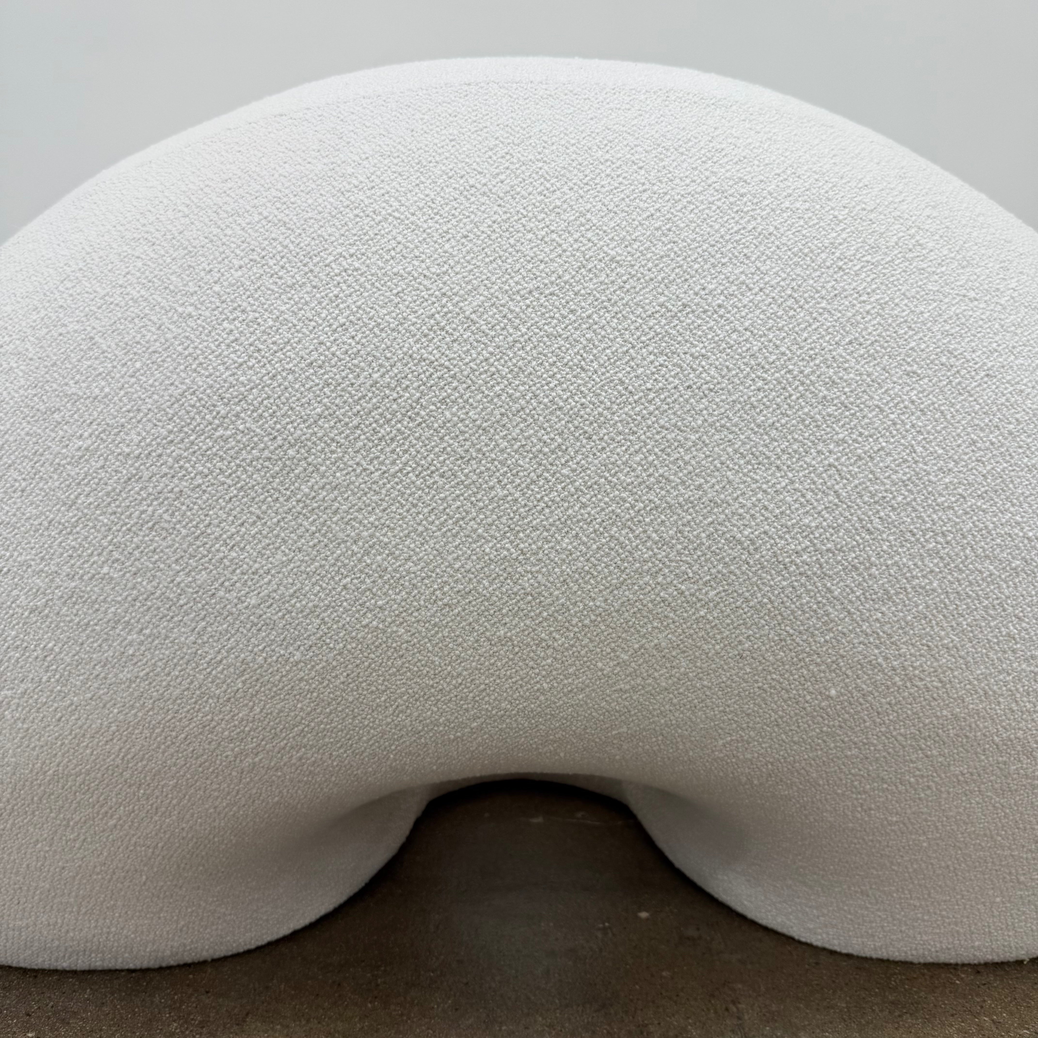 The Objects of Common Interest Tube Chair by Objects of Common Interest is a modern, light grey lounge chair with a donut-shaped design, rounded form, and central indentation, shown against a plain white background.