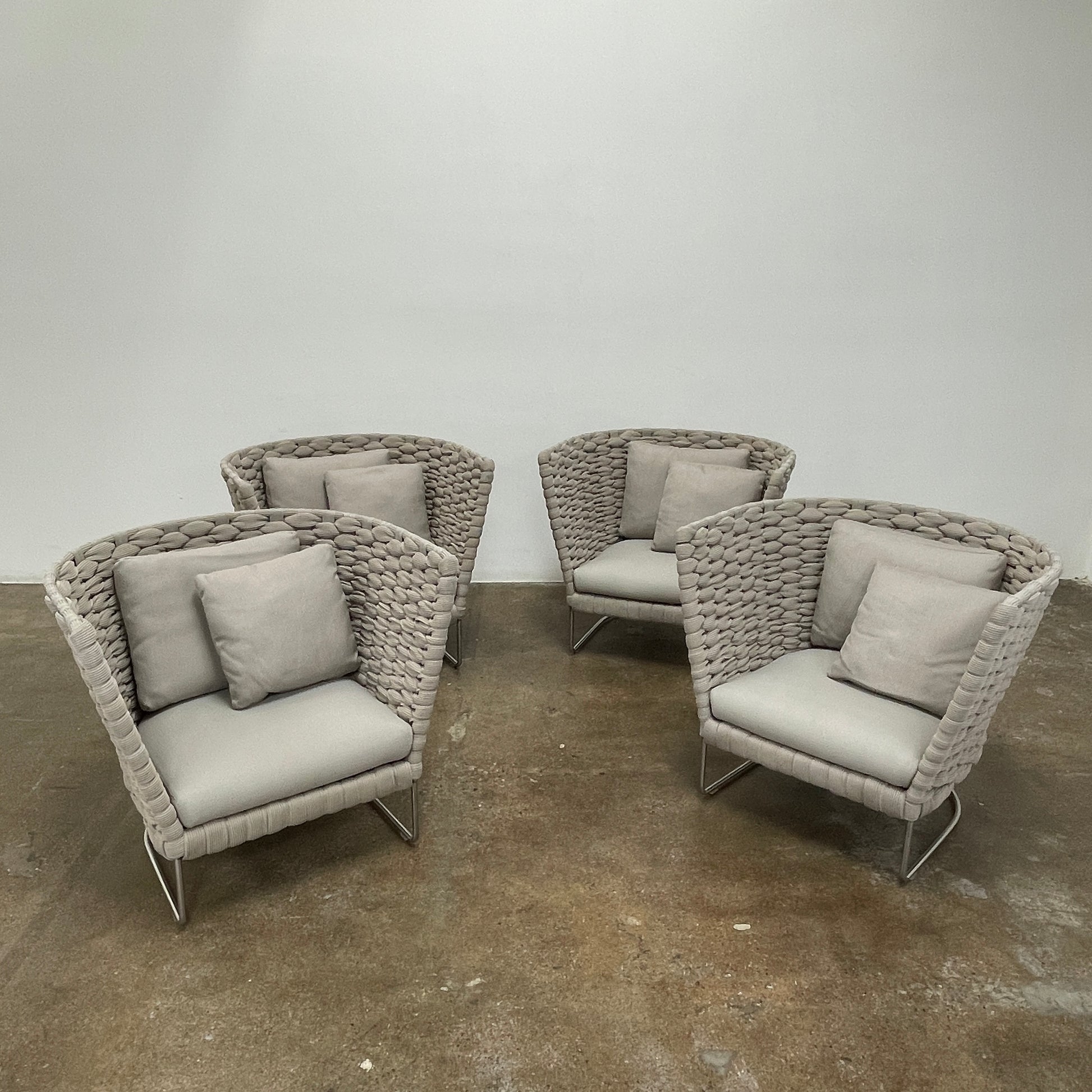 The Paola Lenti Ami Outdoor Armchair by Paola Lenti features a large weave frame, light gray cushions, and a metal base, all set against a plain white background.