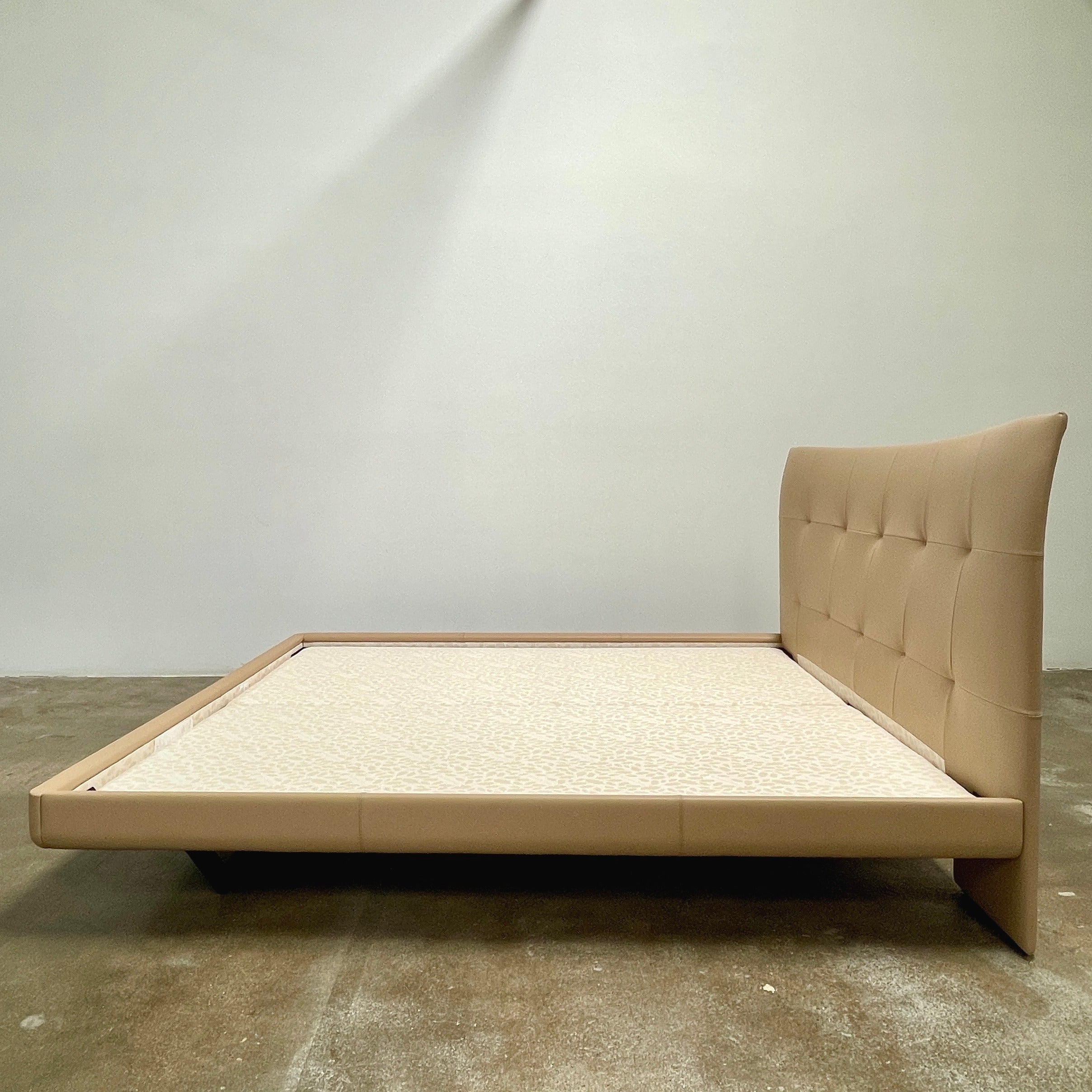 The Poltrona Frau Aurora Due King Bed, crafted by designer Tito Agnoli, boasts a minimalist and modern style with its beige tufted headboard set against a plain light gray wall.