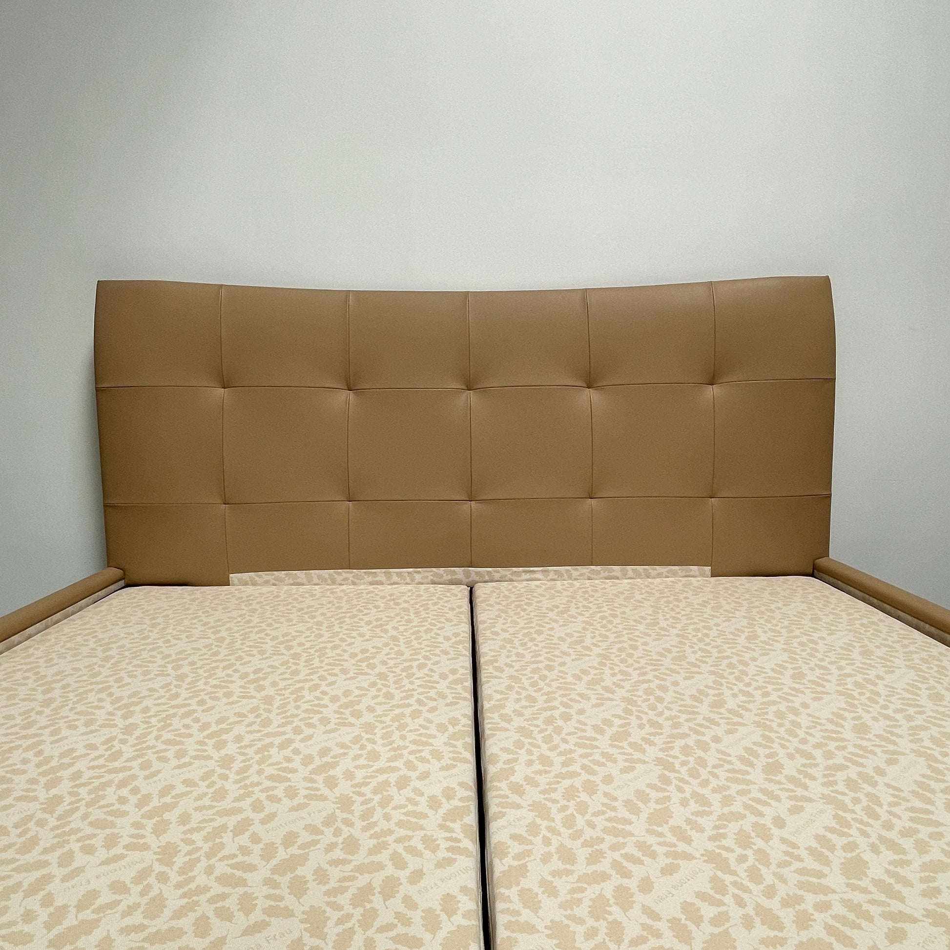 The Poltrona Frau Aurora Due King Bed, crafted by designer Tito Agnoli, boasts a minimalist and modern style with its beige tufted headboard set against a plain light gray wall.