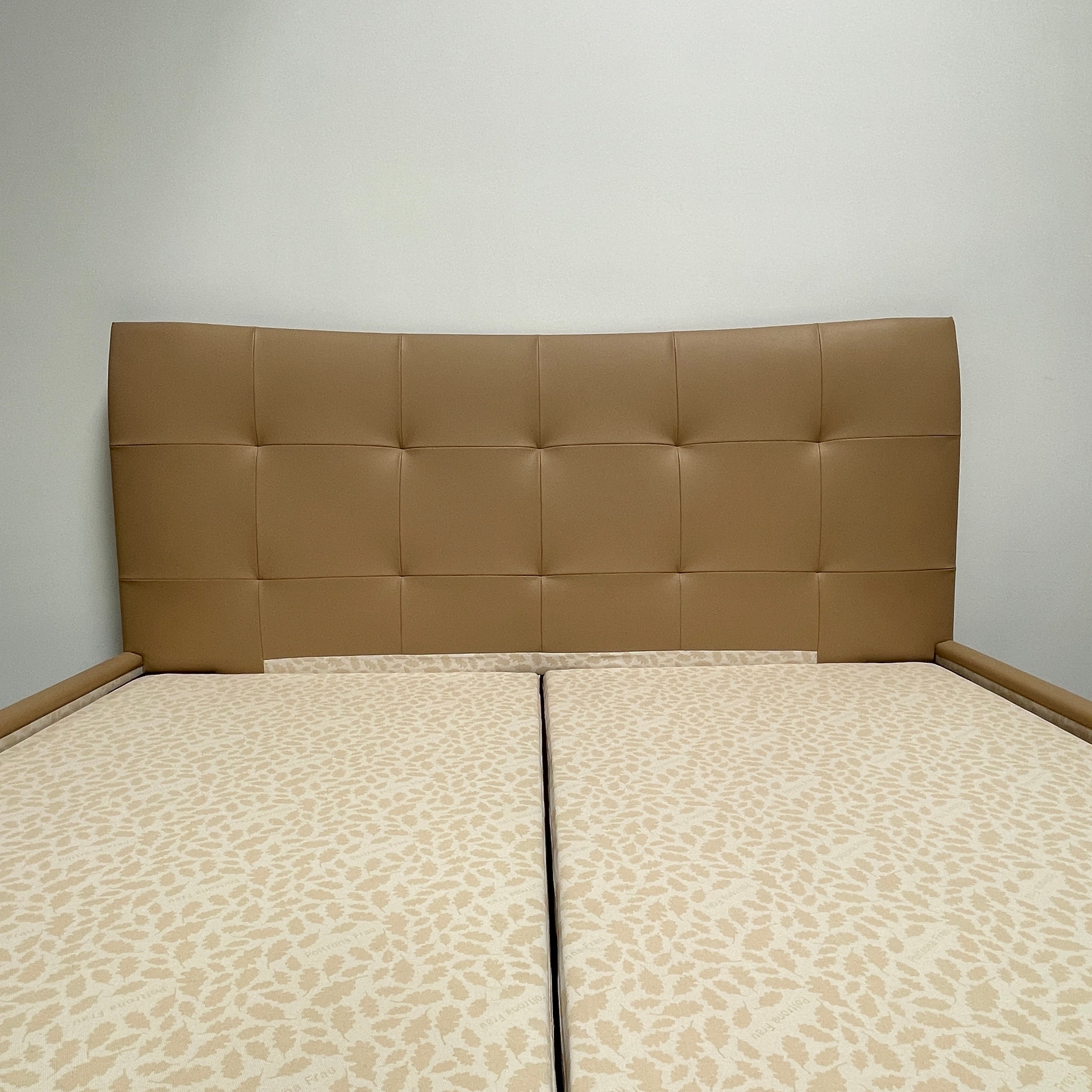 The Poltrona Frau Aurora Due King Bed, crafted by designer Tito Agnoli, boasts a minimalist and modern style with its beige tufted headboard set against a plain light gray wall.