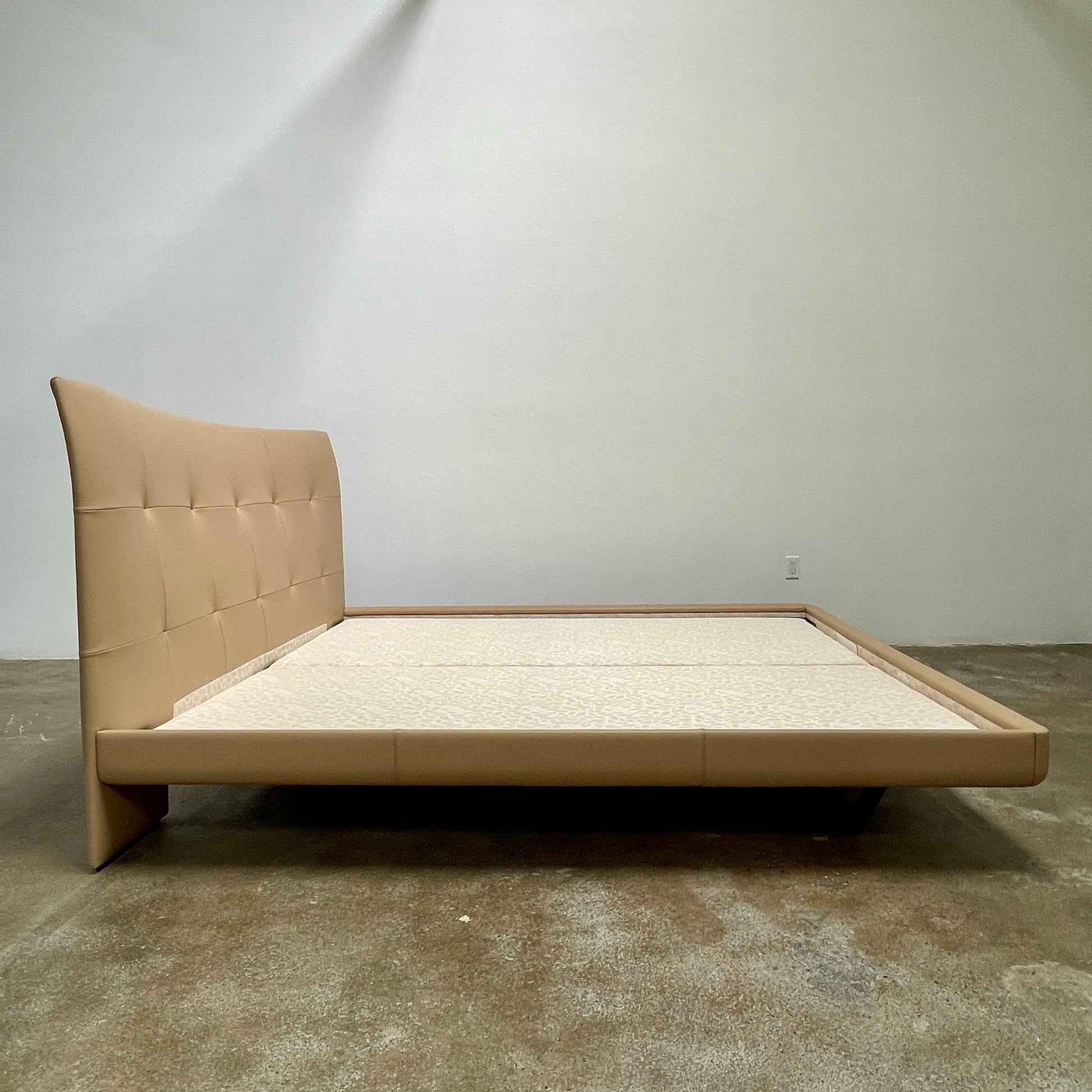 The Poltrona Frau Aurora Due King Bed, crafted by designer Tito Agnoli, boasts a minimalist and modern style with its beige tufted headboard set against a plain light gray wall.