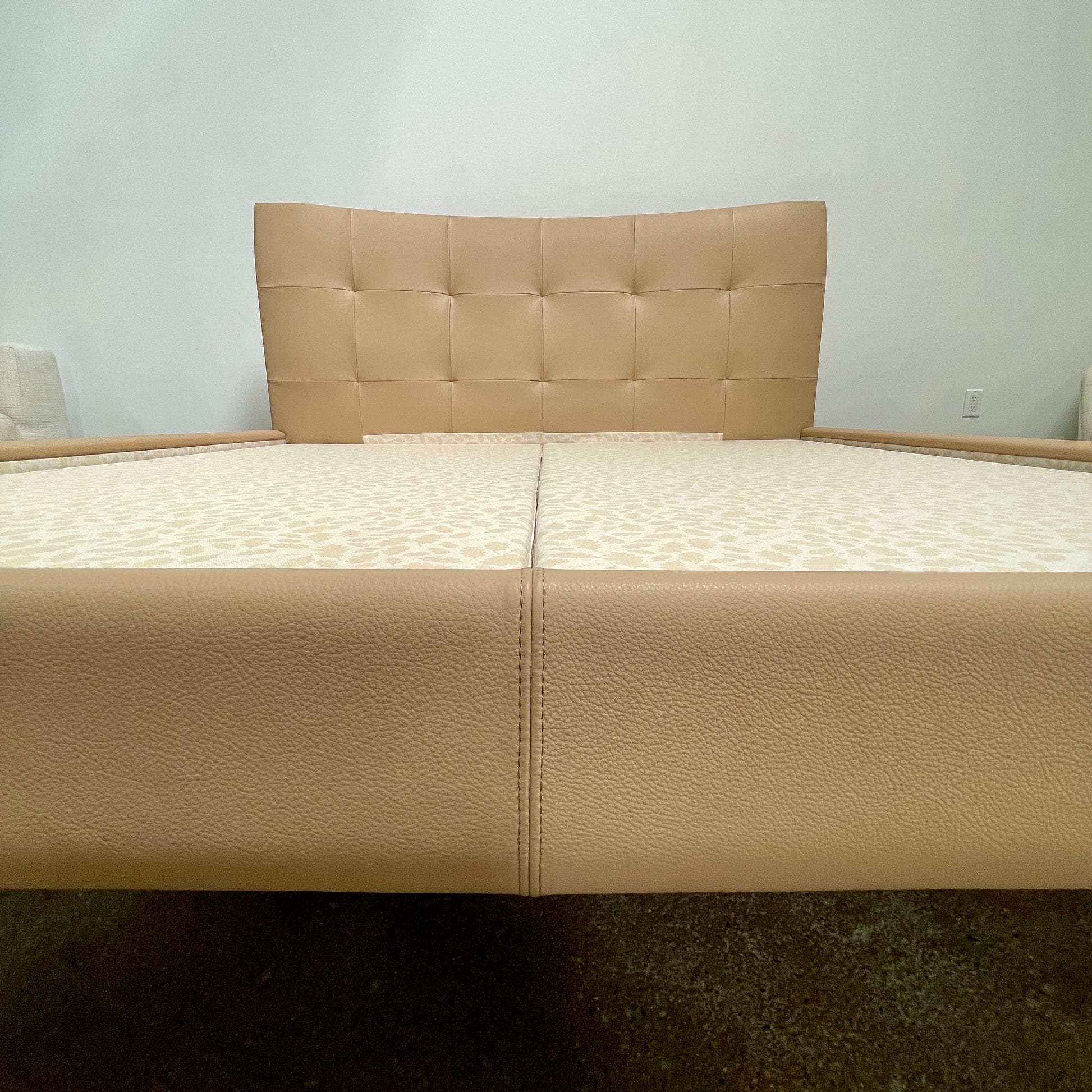 The Poltrona Frau Aurora Due King Bed, crafted by designer Tito Agnoli, boasts a minimalist and modern style with its beige tufted headboard set against a plain light gray wall.