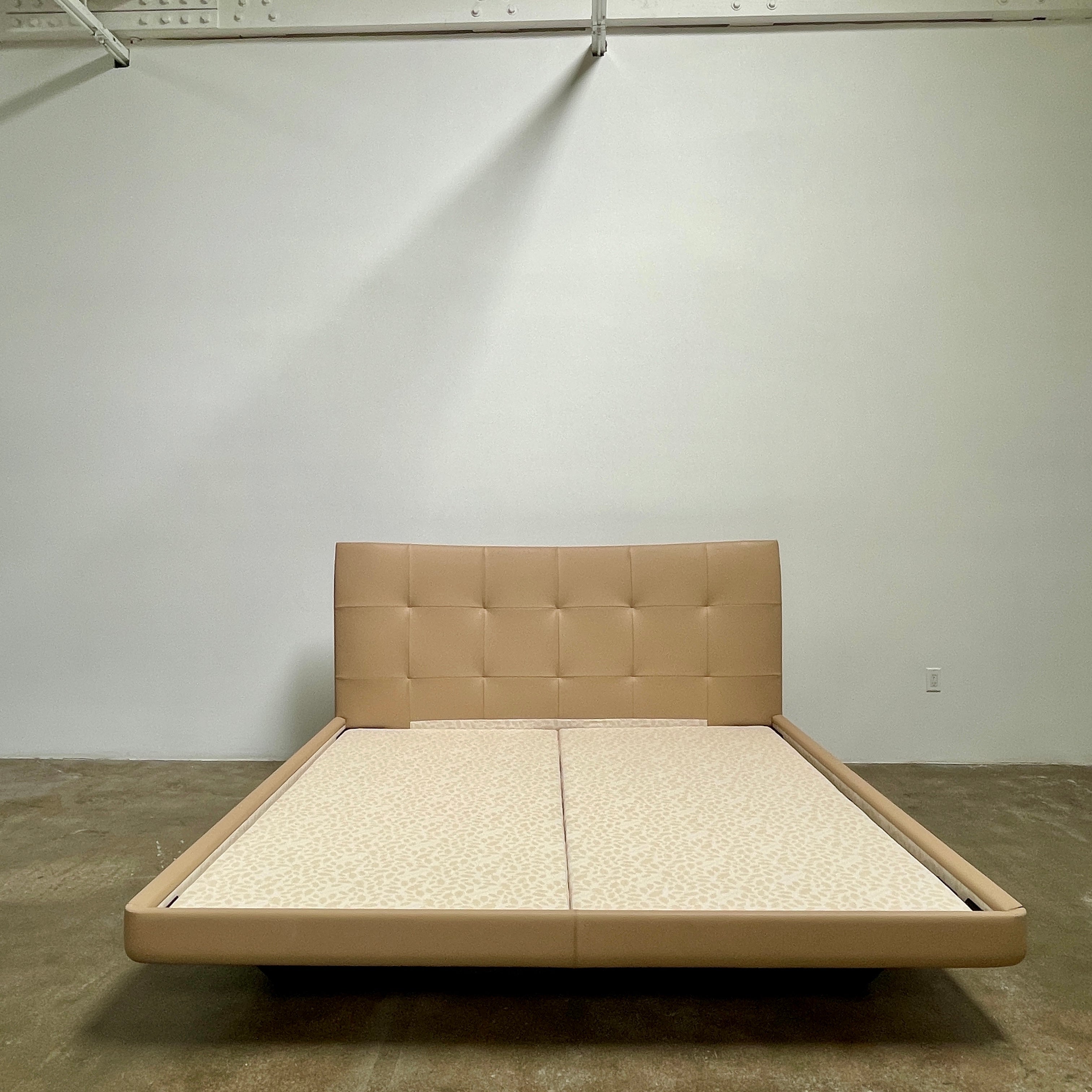 The Poltrona Frau Aurora Due King Bed, crafted by designer Tito Agnoli, boasts a minimalist and modern style with its beige tufted headboard set against a plain light gray wall.