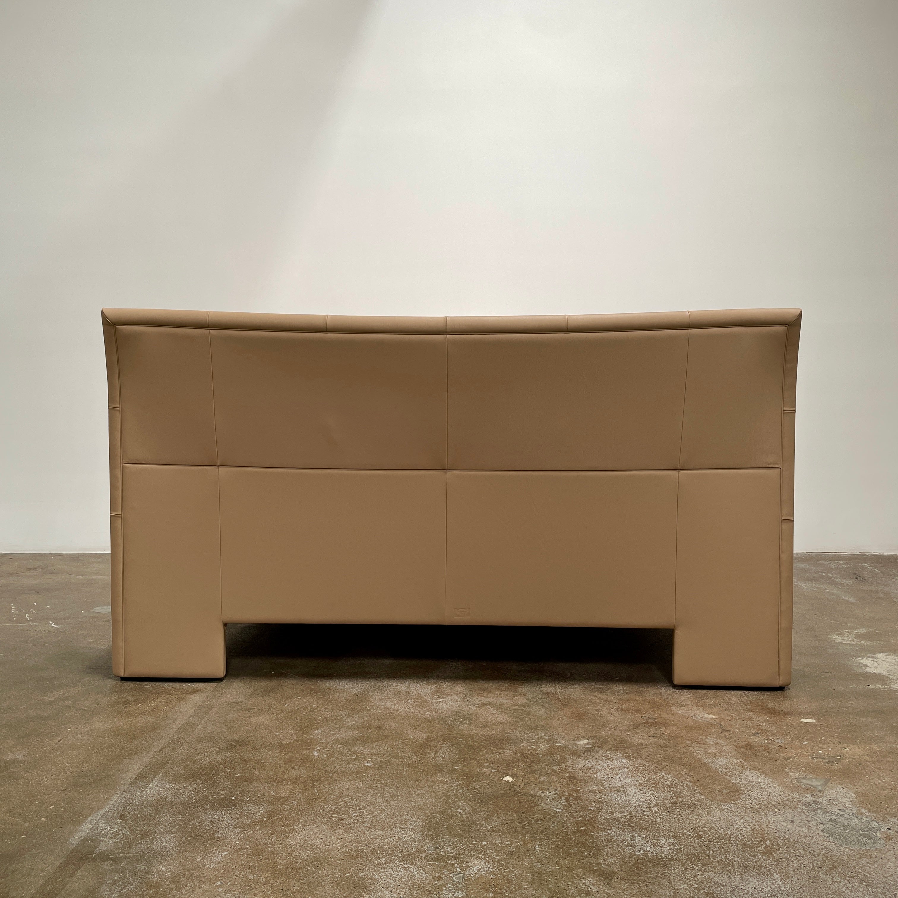 The Poltrona Frau Aurora Due King Bed, crafted by designer Tito Agnoli, boasts a minimalist and modern style with its beige tufted headboard set against a plain light gray wall.