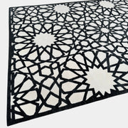 The Roche Bobois Moucharabieh Rug by Roche Bobois features a geometric black and white pattern with interlocking shapes and starbursts, displayed on a plain floor against a white wall.