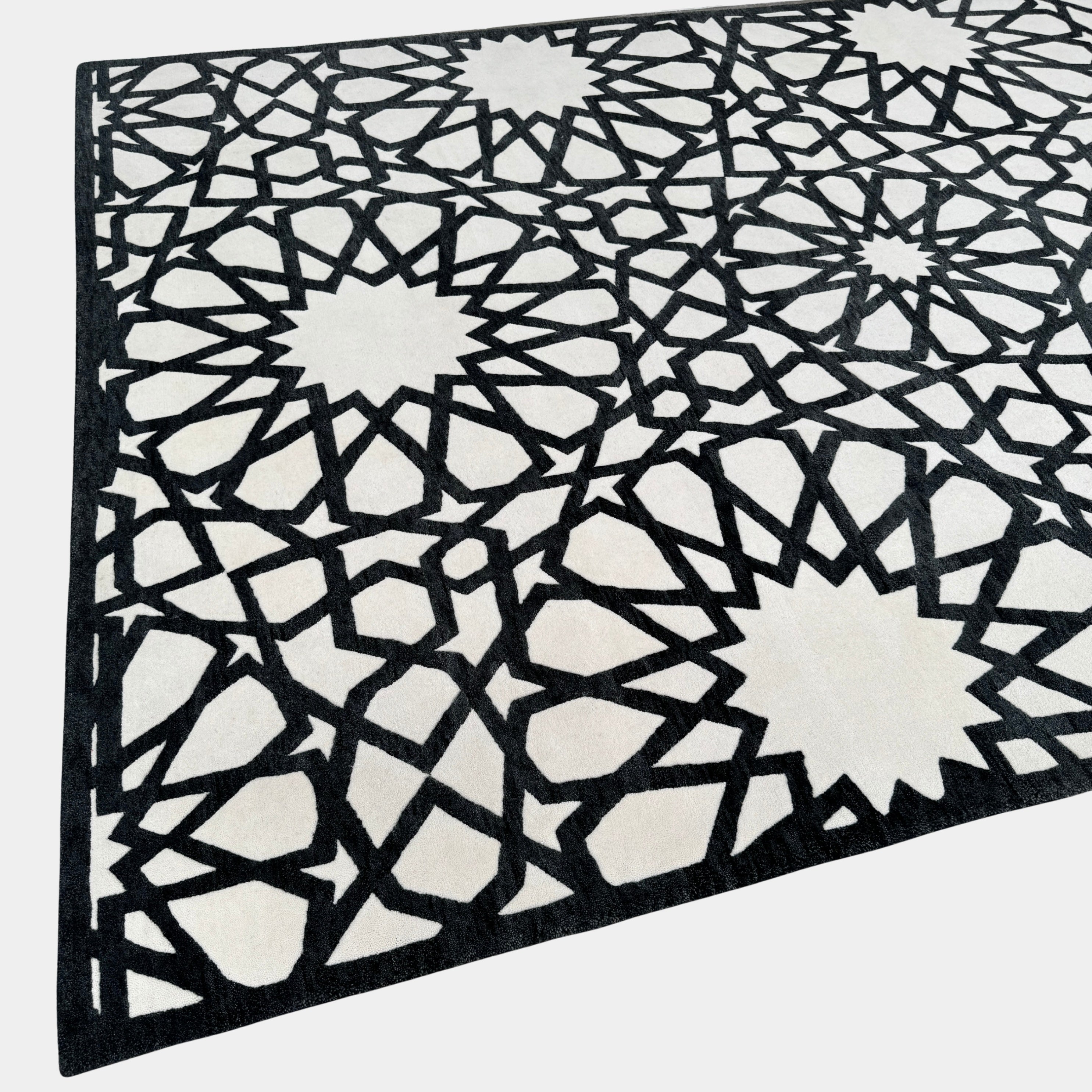 The Roche Bobois Moucharabieh Rug by Roche Bobois features a geometric black and white pattern with interlocking shapes and starbursts, displayed on a plain floor against a white wall.