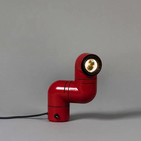 A Santa & Cole Tatu Red Table Lamp with a pipe-shaped design and spotlight head rests on a white surface, plugged in with its black cord.