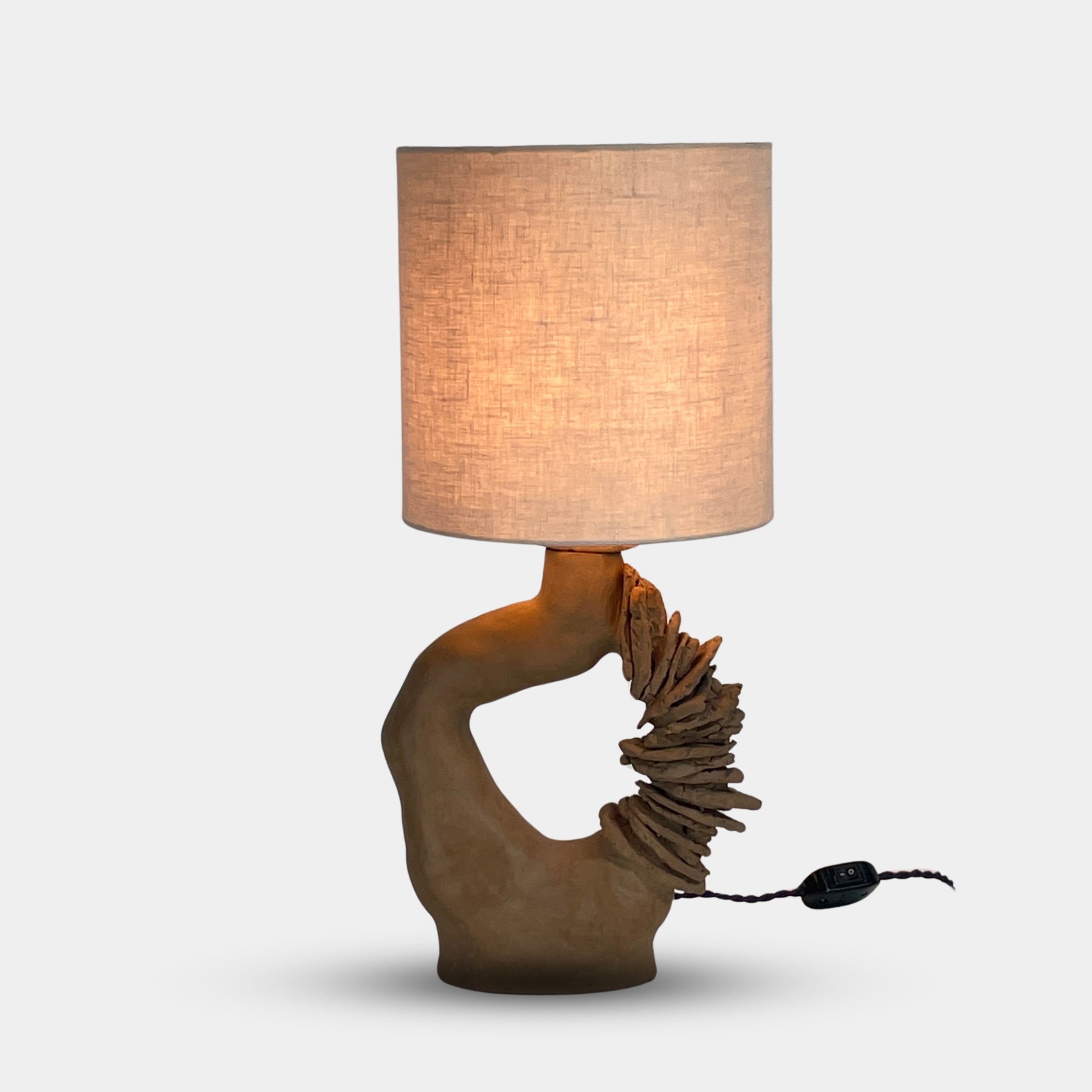 The Sofi Ceramic Arch Lamp by Sofi Ceramics is a distinctively designed table lamp with a twisting sculptural base and a cylindrical shade. Its hand-built ceramic elements softly illuminate against plain backgrounds.