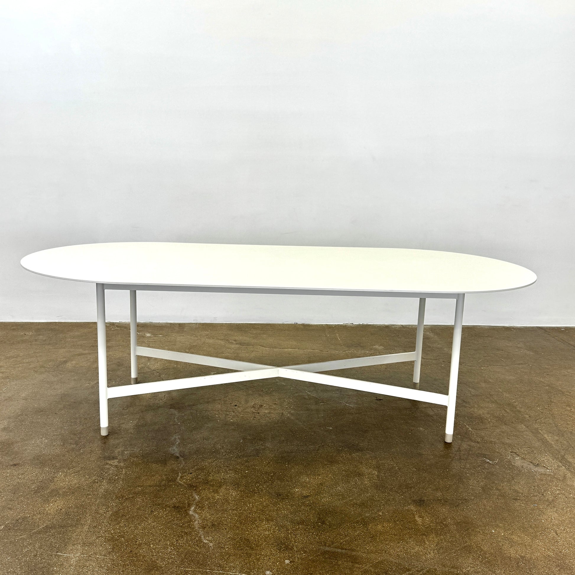 The MillerKnoll Outdoor Sommer Oval Dining Table for DWR, paired with six brown wicker chairs on a polished concrete floor against a white wall, is ideal for fans of modern patio furniture and minimalist design.