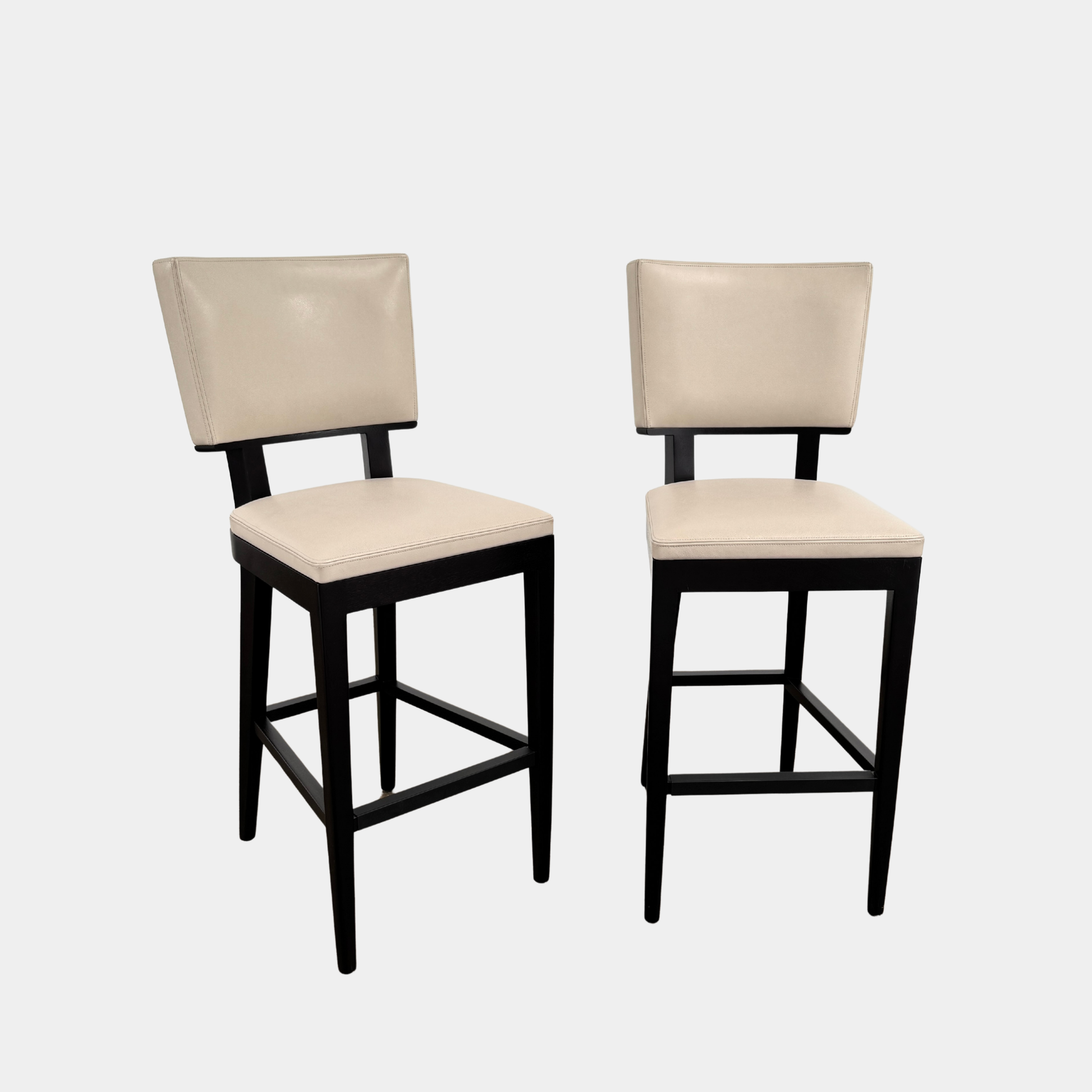 Two Christian Liaigre Bar Stools by Modern Resale, featuring beige upholstery and dark wooden frames, are set on a concrete floor against a white wall, creating a clean and modern branding image.