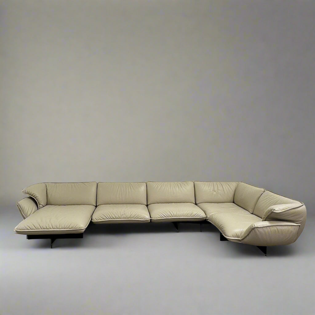 The Cassina Superbeam 5 Piece Leather Sectional in beige, featuring plush cushions, is arranged in a spacious room with white walls, exposed Superbeam ceiling, and overhead lighting.