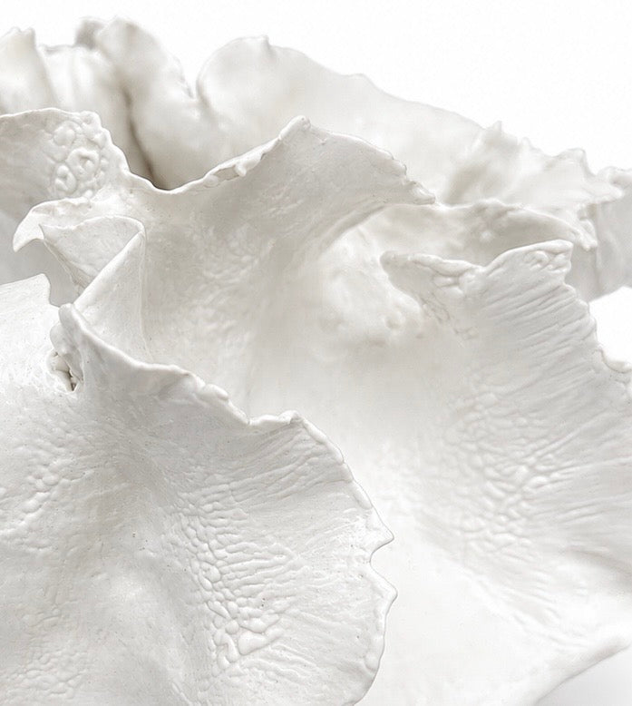 The Sofi Ceramics White Vessel Wave by Artist features ruffled, wavy edges and sits on a square pedestal, highlighting the elegance of handmade pottery against a simple background.