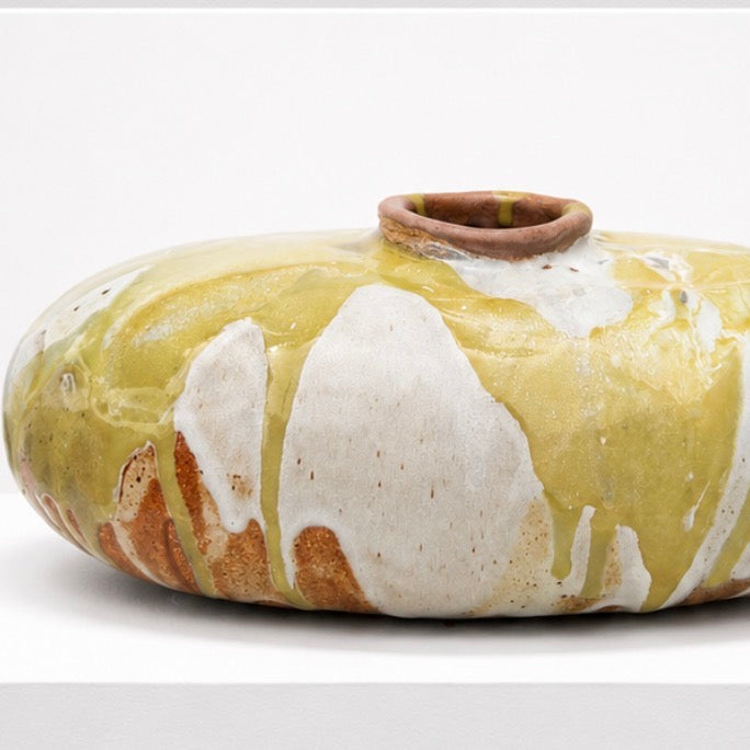 The Sofi Ceramics Marble Vessel by Artist is a round, flat ceramic vase with a small opening, featuring yellow and white glaze drips over a brown base, displayed on a white surface.