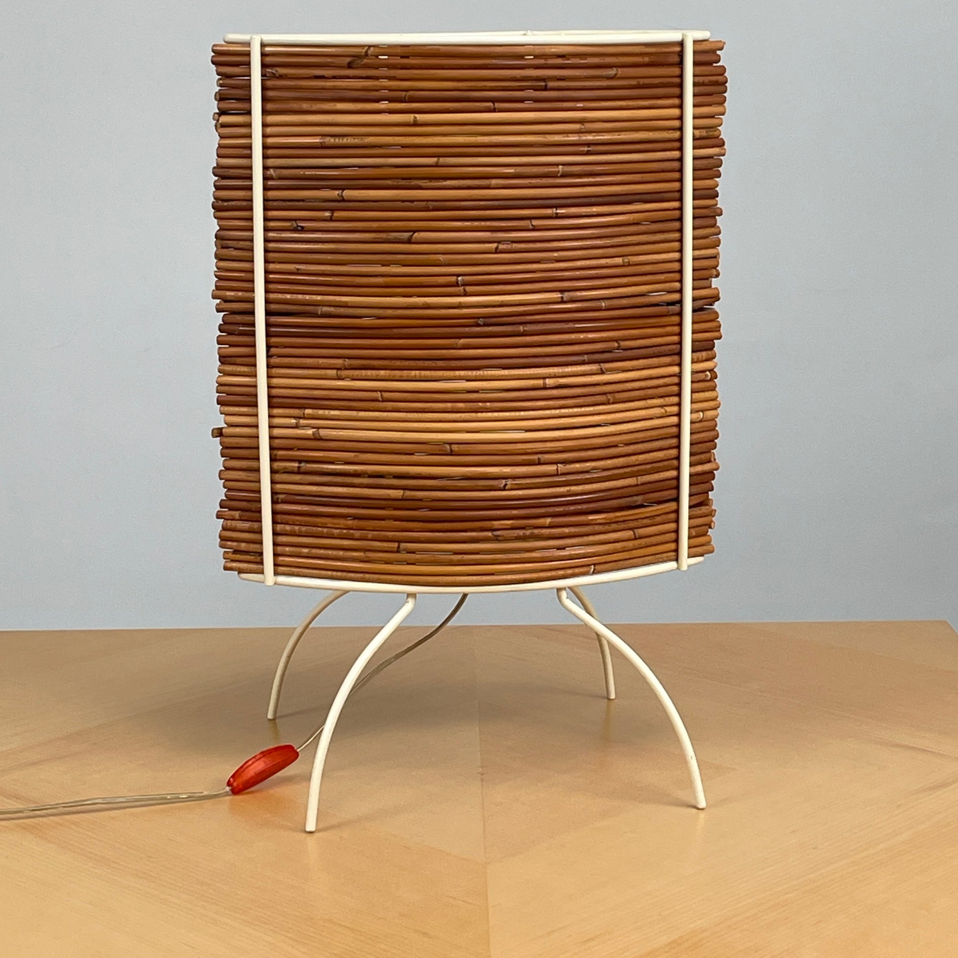 The Vintage Fontana Arte Bambú Table Light by Fontana Arte showcases a woven rattan circular shade atop a sleek white metal tripod base, making it an elegant addition to a wooden table. This lamp enhances your home's interior lighting with its natural charm and unique design.