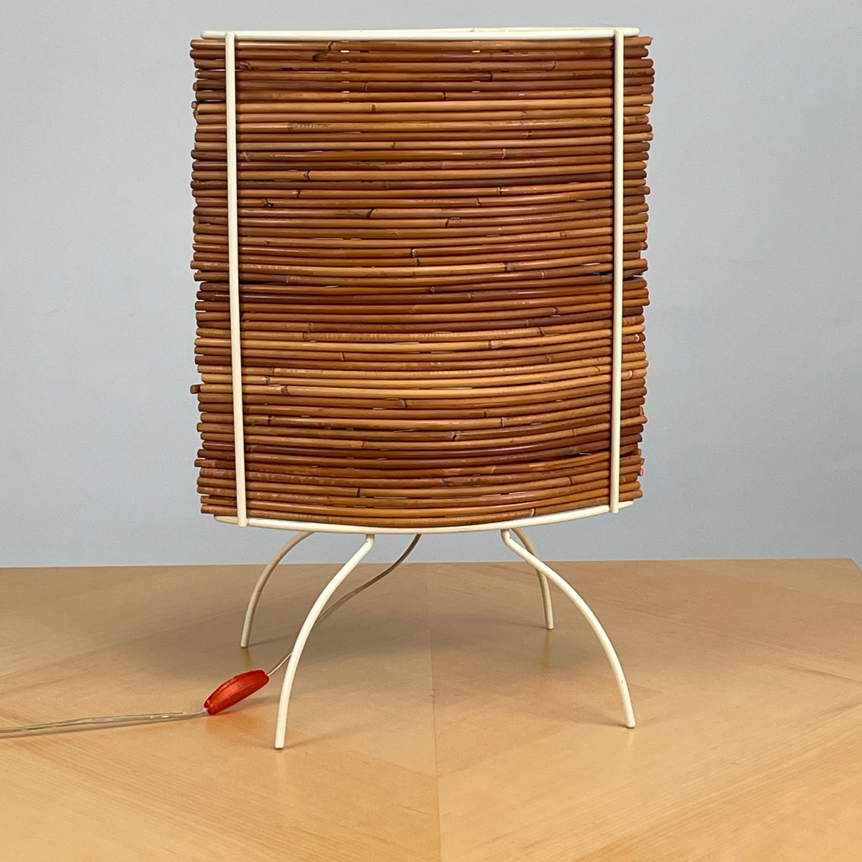 The Vintage Fontana Arte Bambú Table Light by Fontana Arte showcases a woven rattan circular shade atop a sleek white metal tripod base, making it an elegant addition to a wooden table. This lamp enhances your home's interior lighting with its natural charm and unique design.