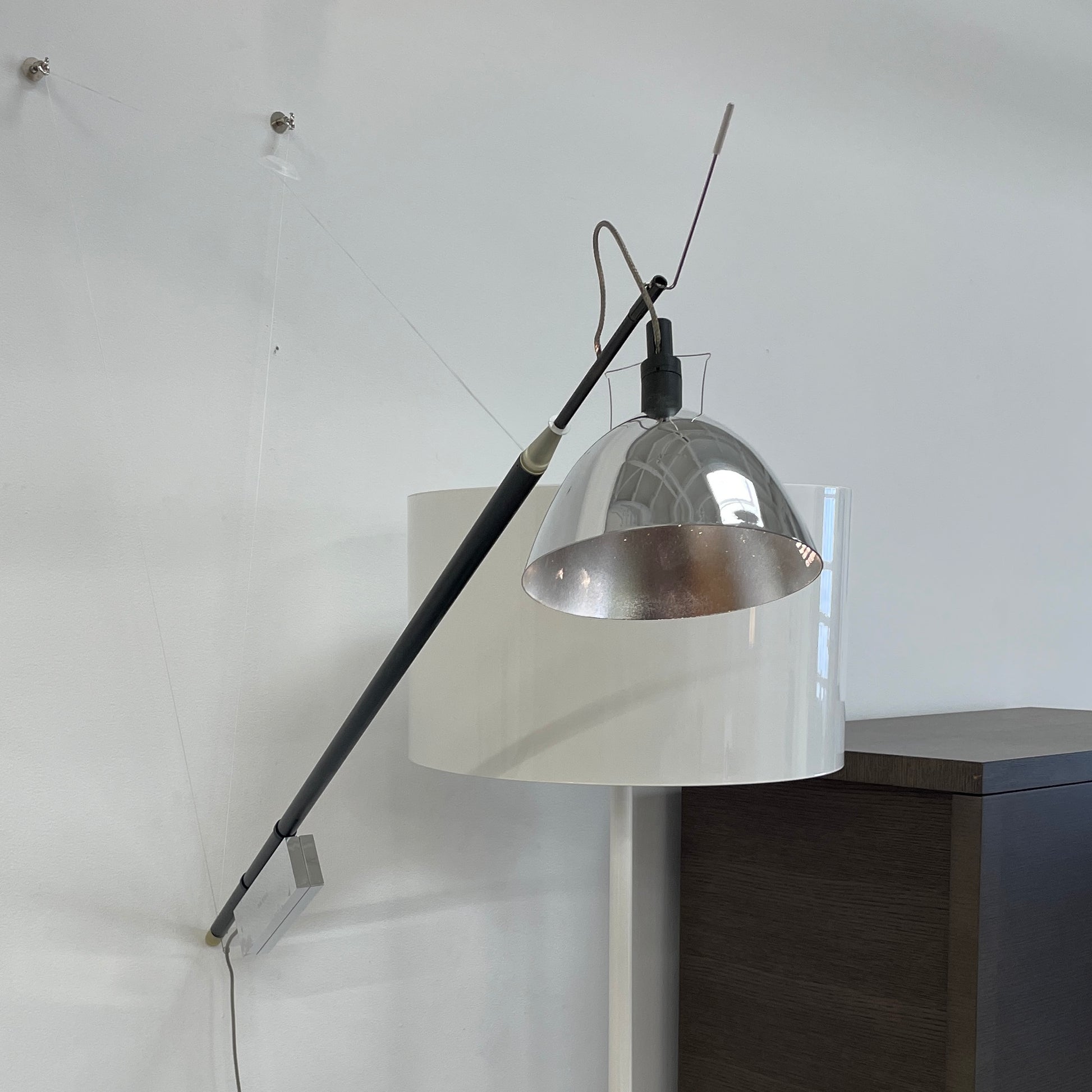 The Ingo Maurer Max Mover Wall Light by Ingo Maurer is a contemporary fixture with an adjustable telescopic rod, sleek black arm, and aluminum reflector in a dome-shaped shade set against a light grey backdrop.
