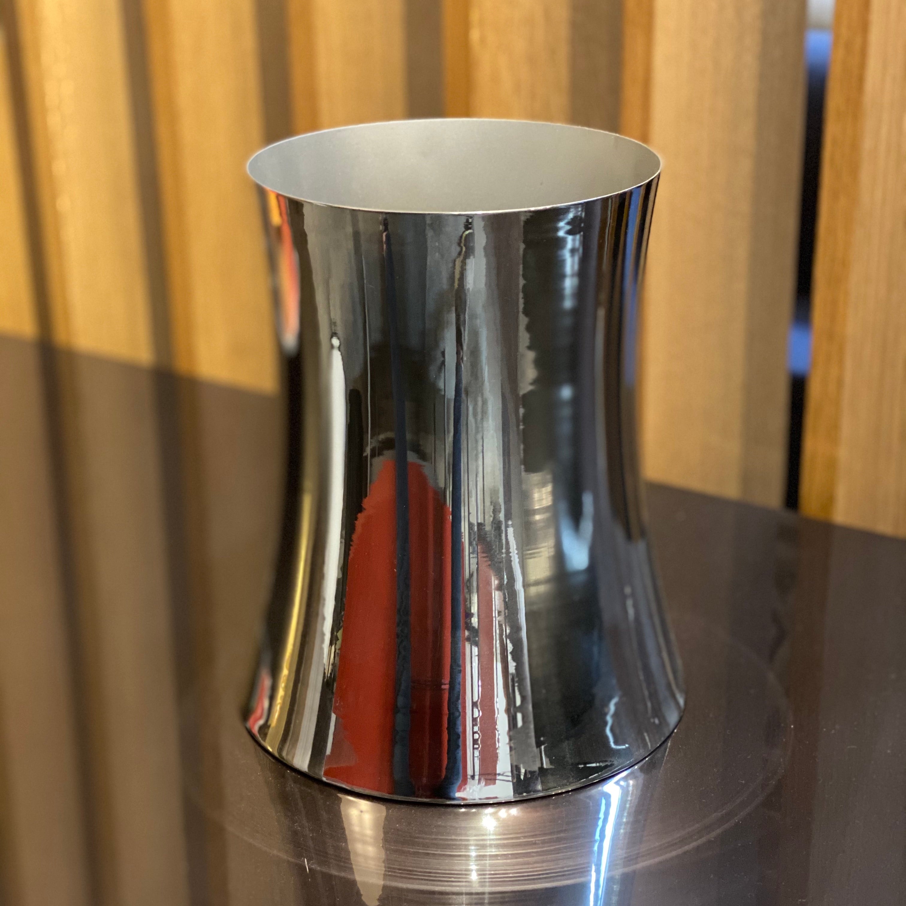 The Zubazuba Vessel by Leo Aerts, from Alinea Design Objects, is crafted from polished stainless steel with a subtly concave middle design. It stands elegantly upright against a simple backdrop, showcasing its versatility as both an artistic piece and a sophisticated champagne bucket.