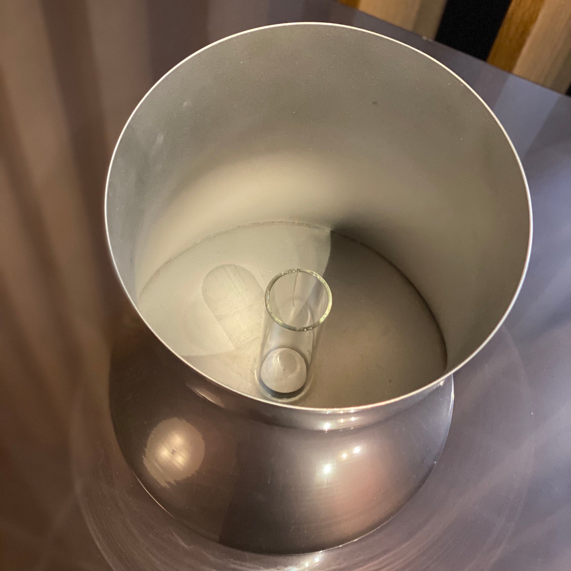 The Zubazuba Vessel by Leo Aerts, from Alinea Design Objects, is crafted from polished stainless steel with a subtly concave middle design. It stands elegantly upright against a simple backdrop, showcasing its versatility as both an artistic piece and a sophisticated champagne bucket.