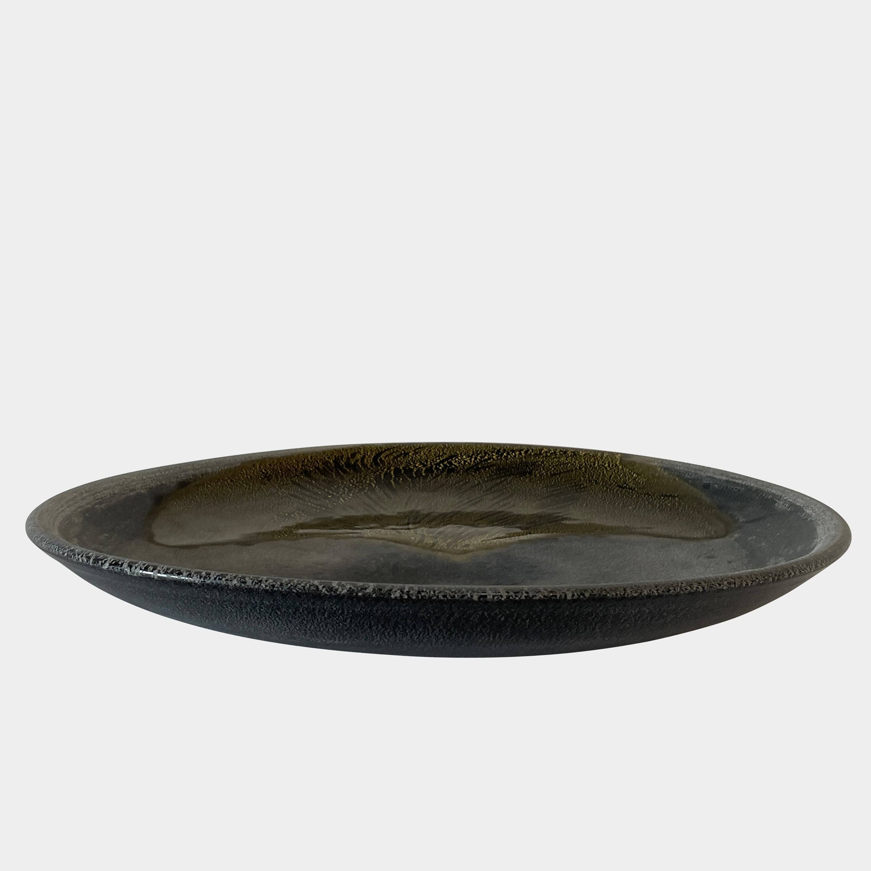 A round ceramic plate featuring a central raised circular design, predominantly gray with earthy bronze tones and textured streaks on one side, reminiscent of the artistry seen in the Alfredo Barbini Murano Glass Plate And Object.