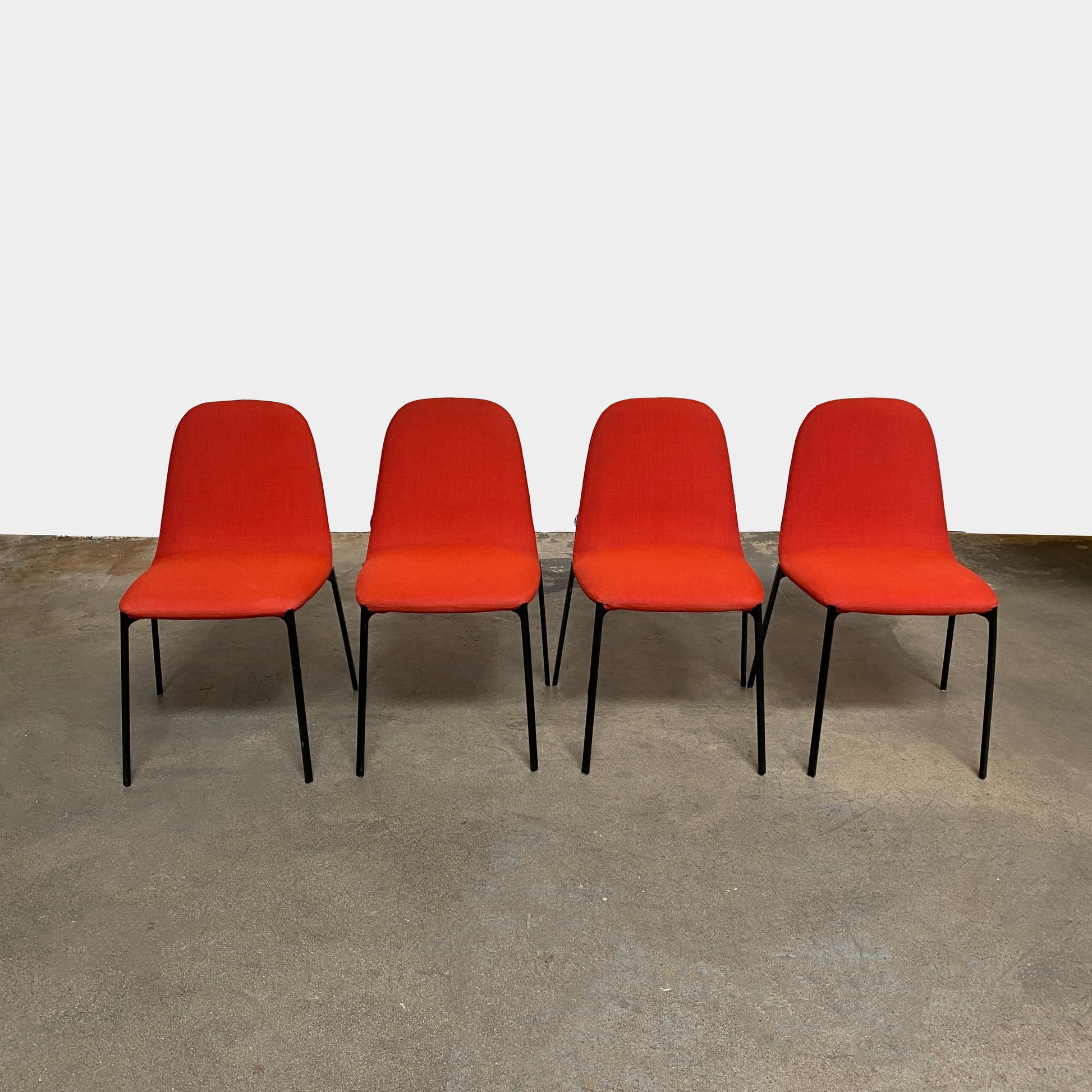 Four Zanotta Ella Dining Chairs, with their red fabric and black metal legs, are set in a row against a plain white background, showcasing the sleek design of this Italian brand.
