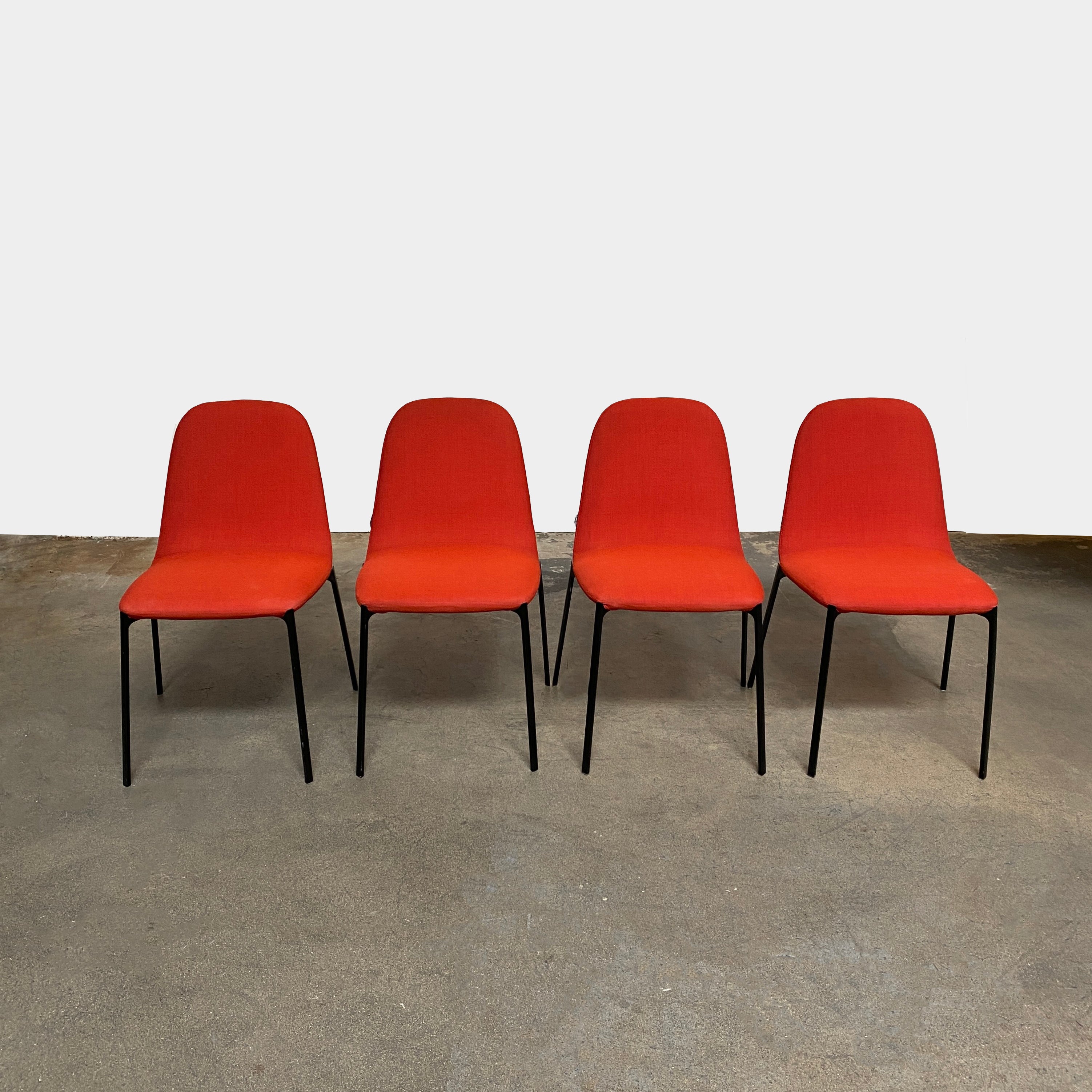 Four Zanotta Ella Dining Chairs, with their red fabric and black metal legs, are set in a row against a plain white background, showcasing the sleek design of this Italian brand.