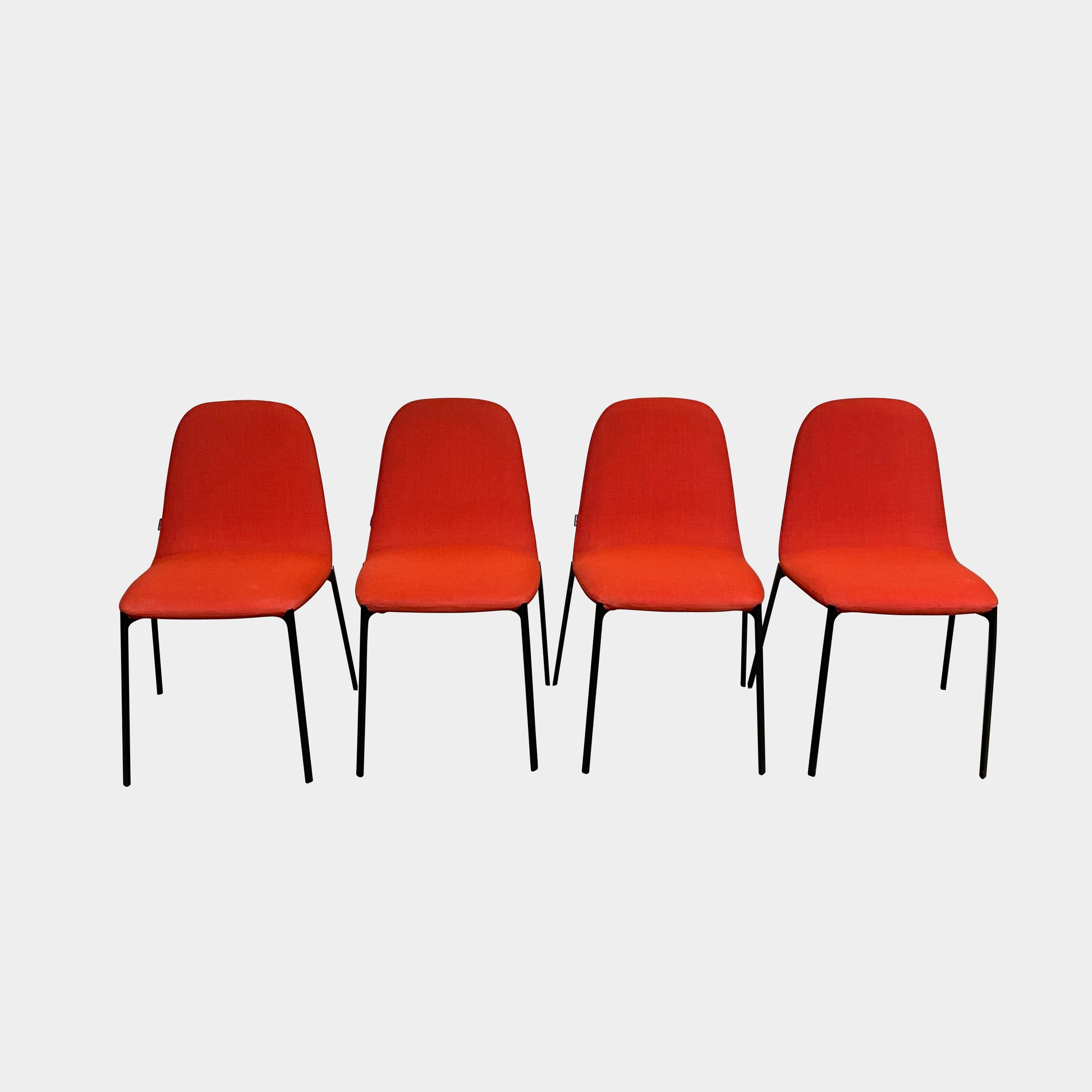 Four Zanotta Ella Dining Chairs, with their red fabric and black metal legs, are set in a row against a plain white background, showcasing the sleek design of this Italian brand.