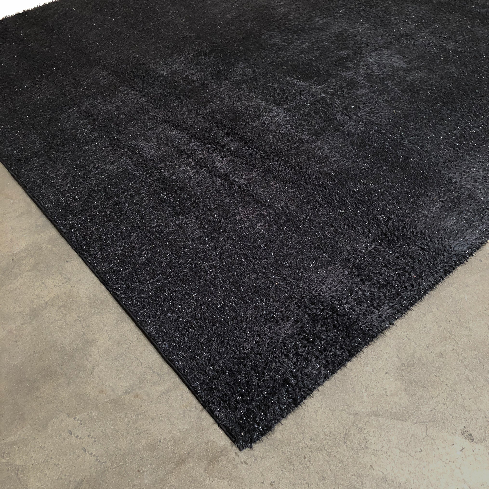 A close-up of the corner of a Kasthall Monroe Speckled Shag Rug on a plain, light-colored floor.