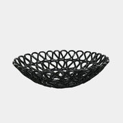 A Tom Dixon Fresh Fat Bowl in black with an intricate weave rests on a concrete floor, set against a simple white backdrop.